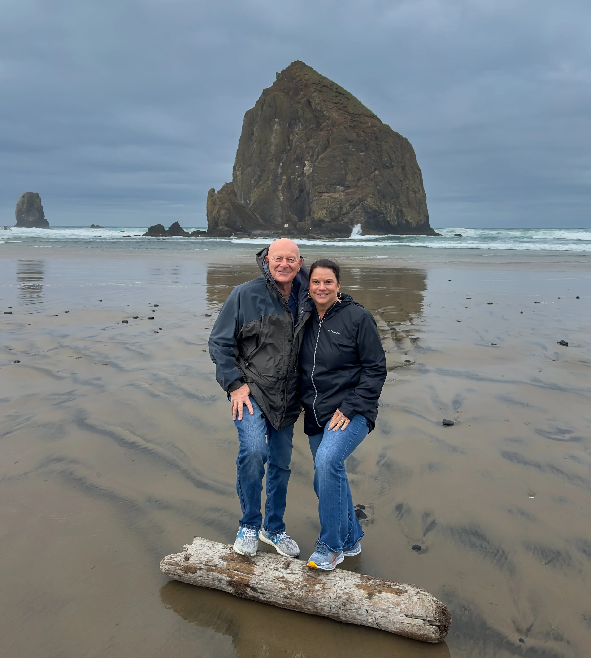 Two people standing on a piece of driftwood on a beach with large sea stacks in the background. Overcast sky.