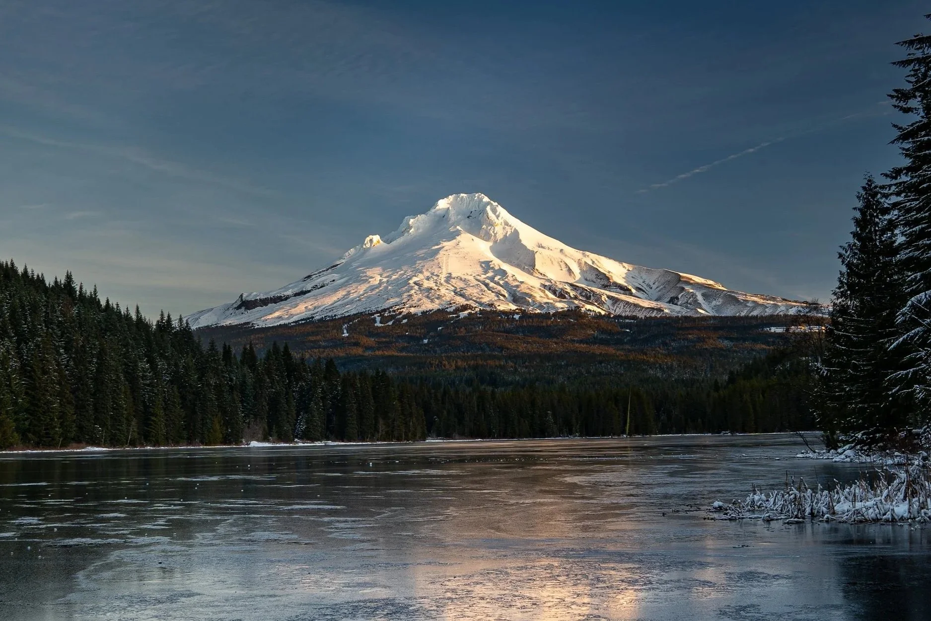 Snow-covered mountain behind a forest, with a partially frozen lake in the foreground, under a clear sky.
