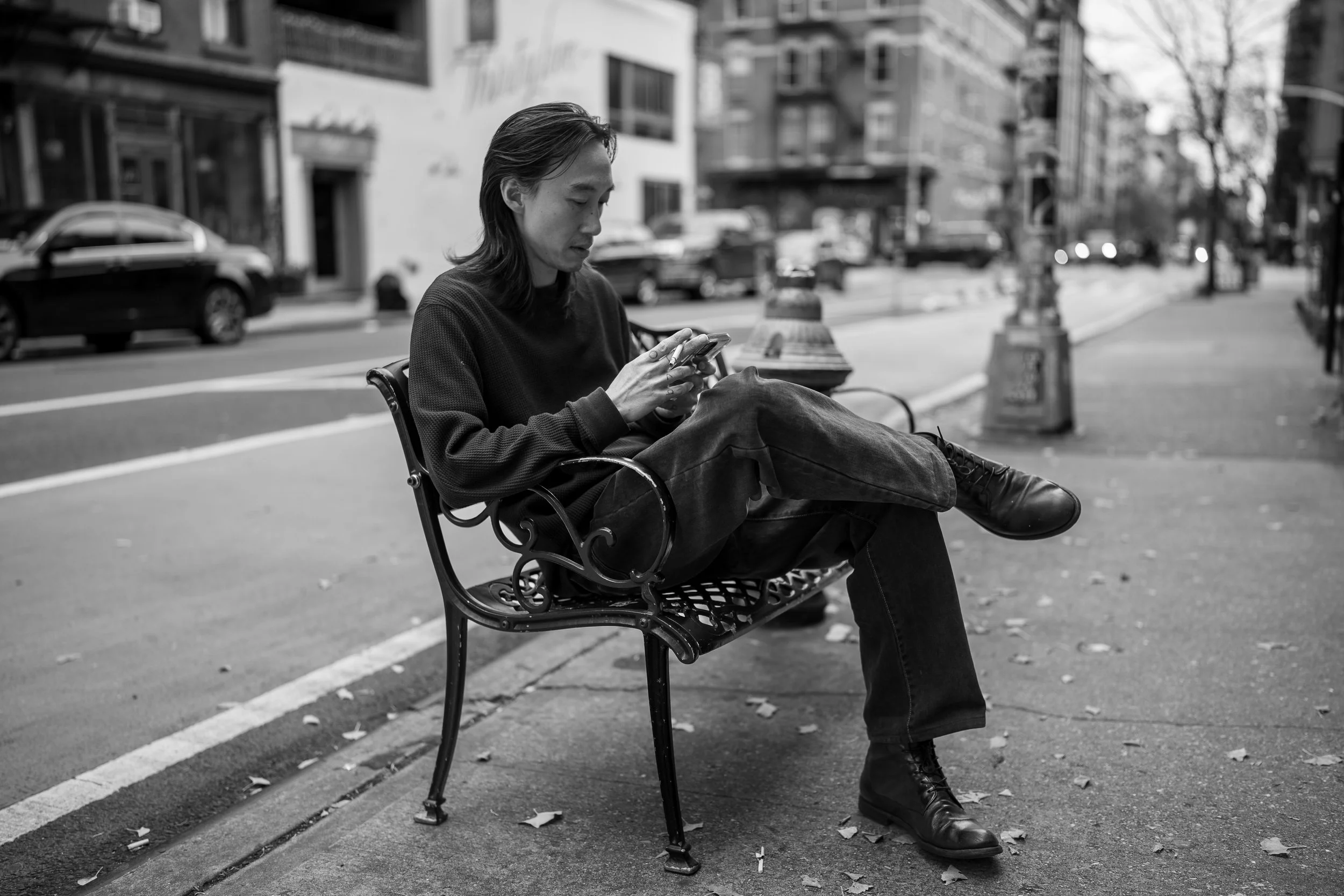 A man sitting on a NYC bench in an urban setting, looking at his phone. Black-and-white photo with cars and buildings in the background.