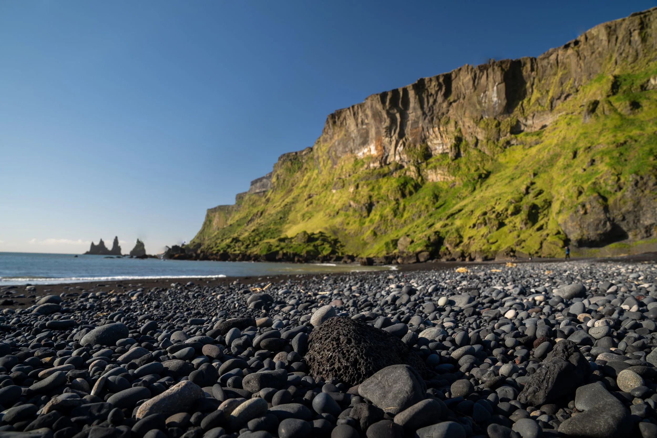 A rocky beach with smooth gray stones leading to a green cliffside and the ocean, with two sea stacks in the distance and a person walking along the shoreline.