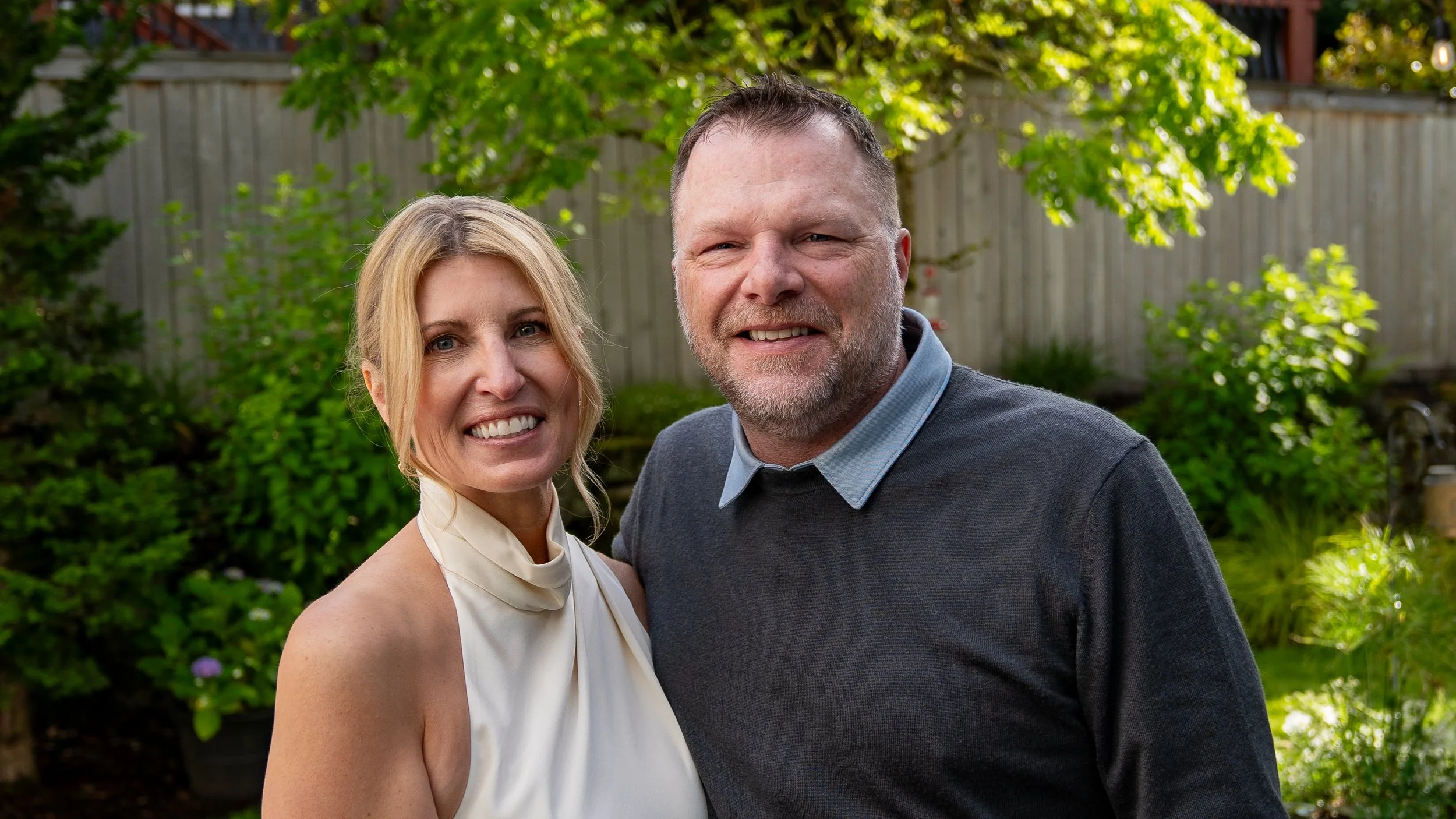 A smiling couple standing outdoors in a garden with green trees and bushes in the background, sunlight filtering through the leaves.