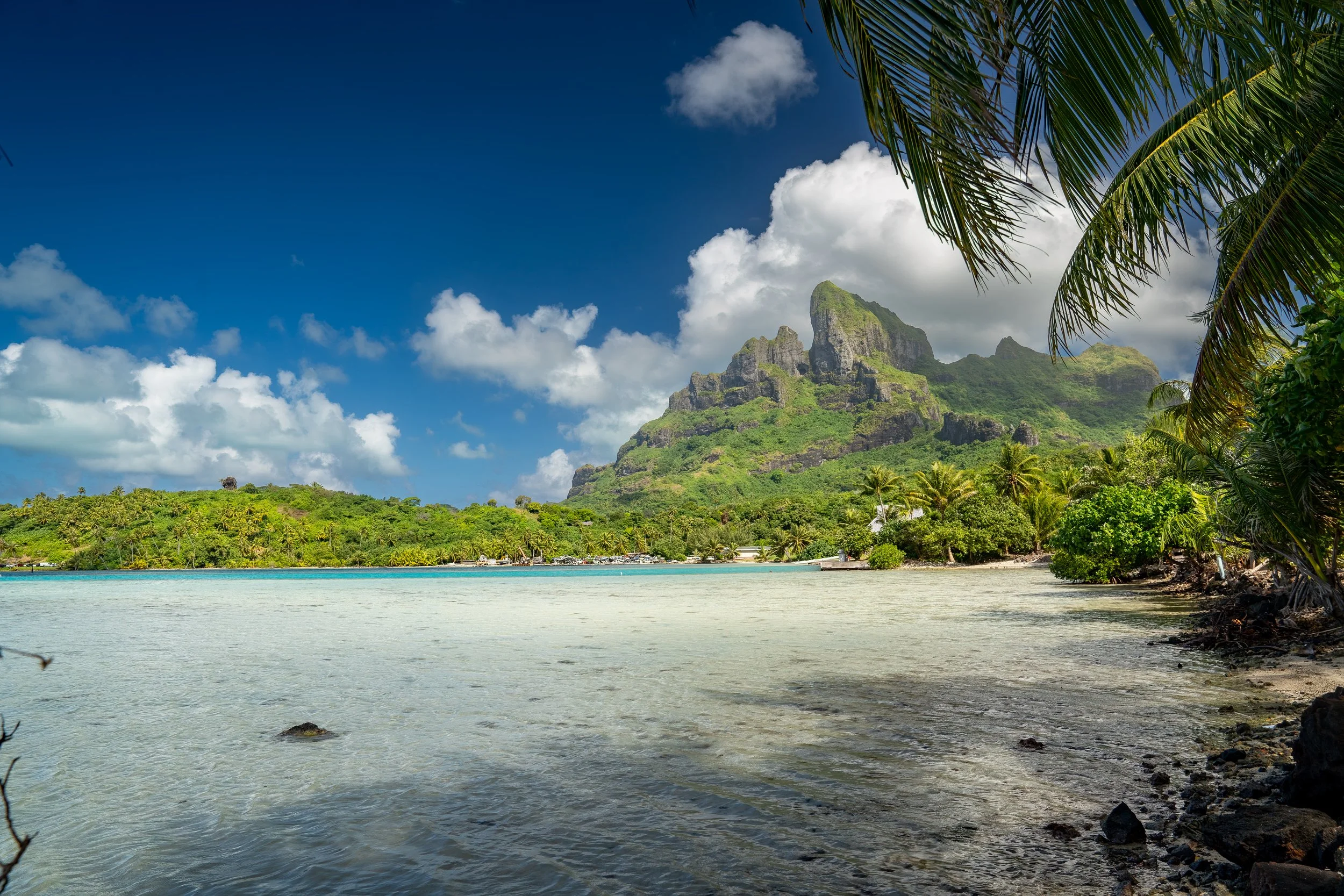 Bora Bora is a French Island group in the Leeward Islands. Tropical island with lush green mountains, clear blue water, and palm trees, under a partly cloudy sky.