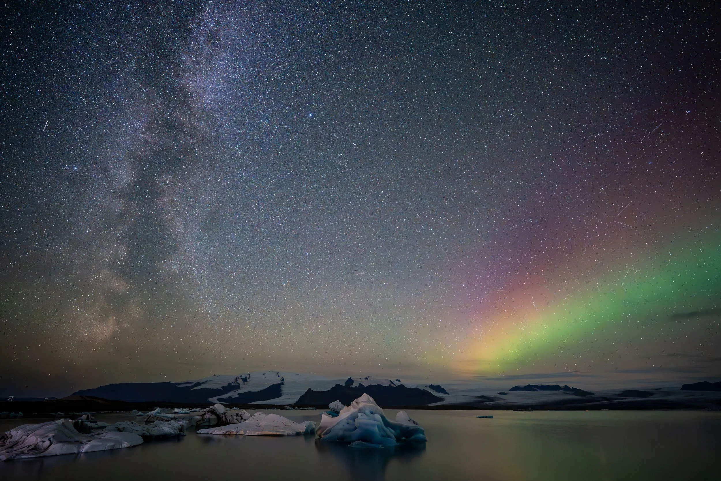 Night sky filled with stars and the Milky Way galaxy, with a faint green aurora at the horizon. In the foreground, icy landscape with floating ice formations on calm water and snow-covered mountains in the distance.