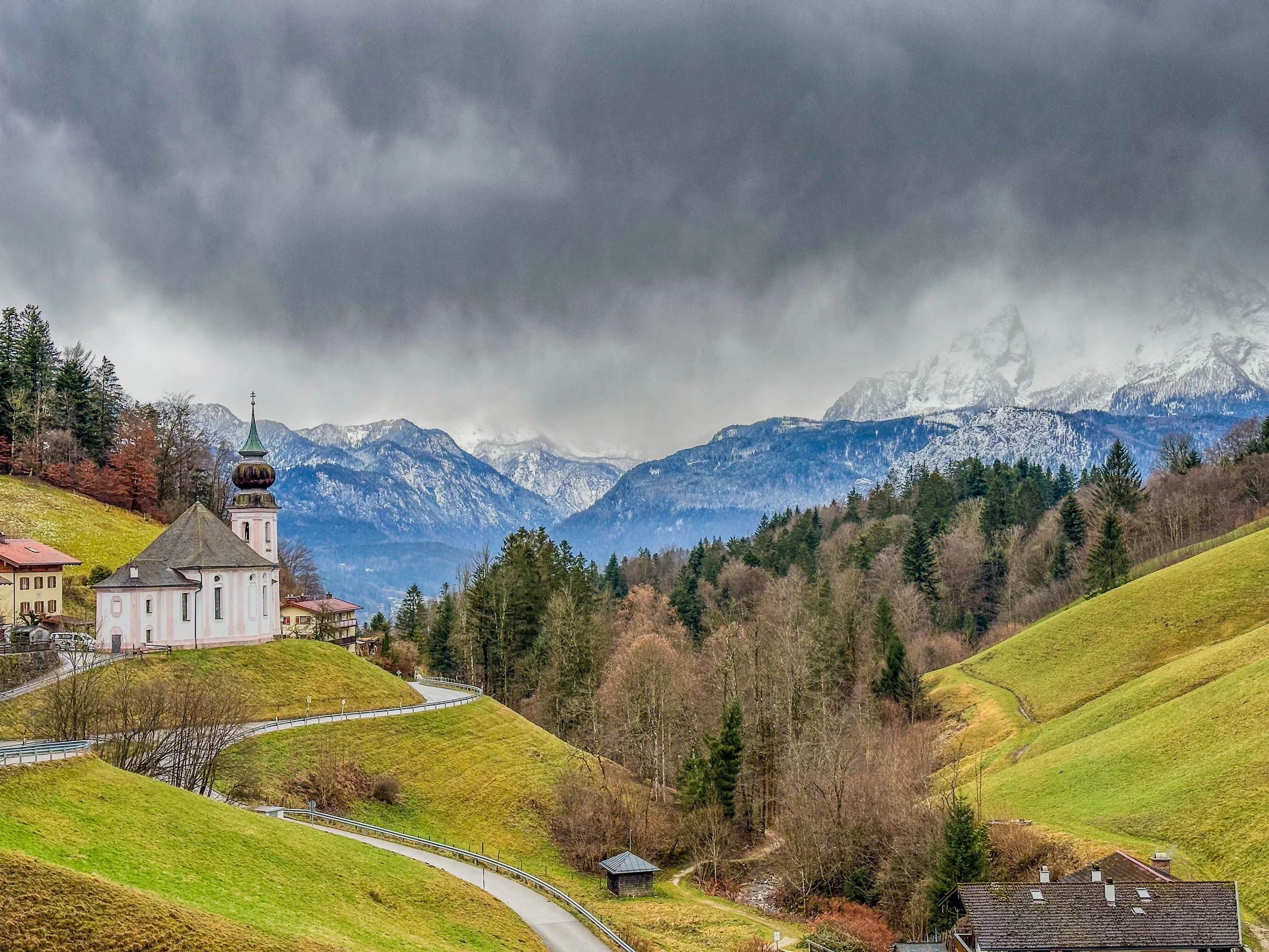 Scenic landscape with green rolling hills, a winding road, a church with a green steeple, forested areas, snow-capped mountains, and dark, cloudy sky.