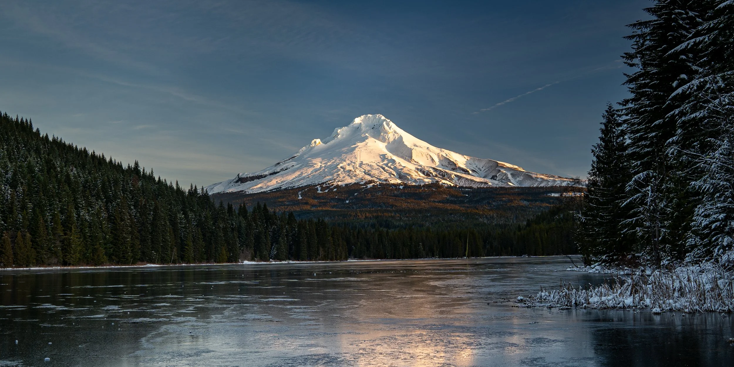 A snow-capped mountain, likely Mount Hood, viewed from across a frozen lake with pine trees on either side.
