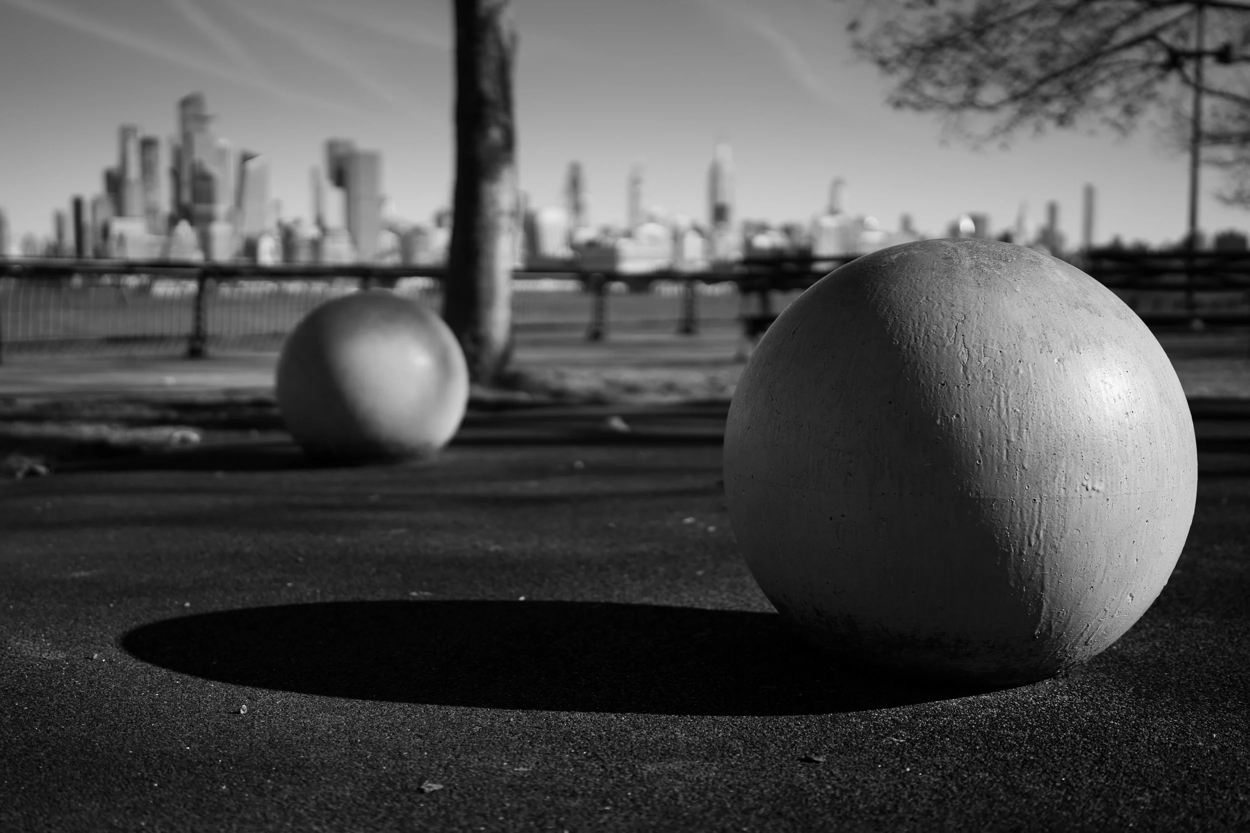 Black and white outdoor scene with two large spherical objects casting shadows on the ground, a wooden bench, trees with bare branches, and a city skyline in the background.
