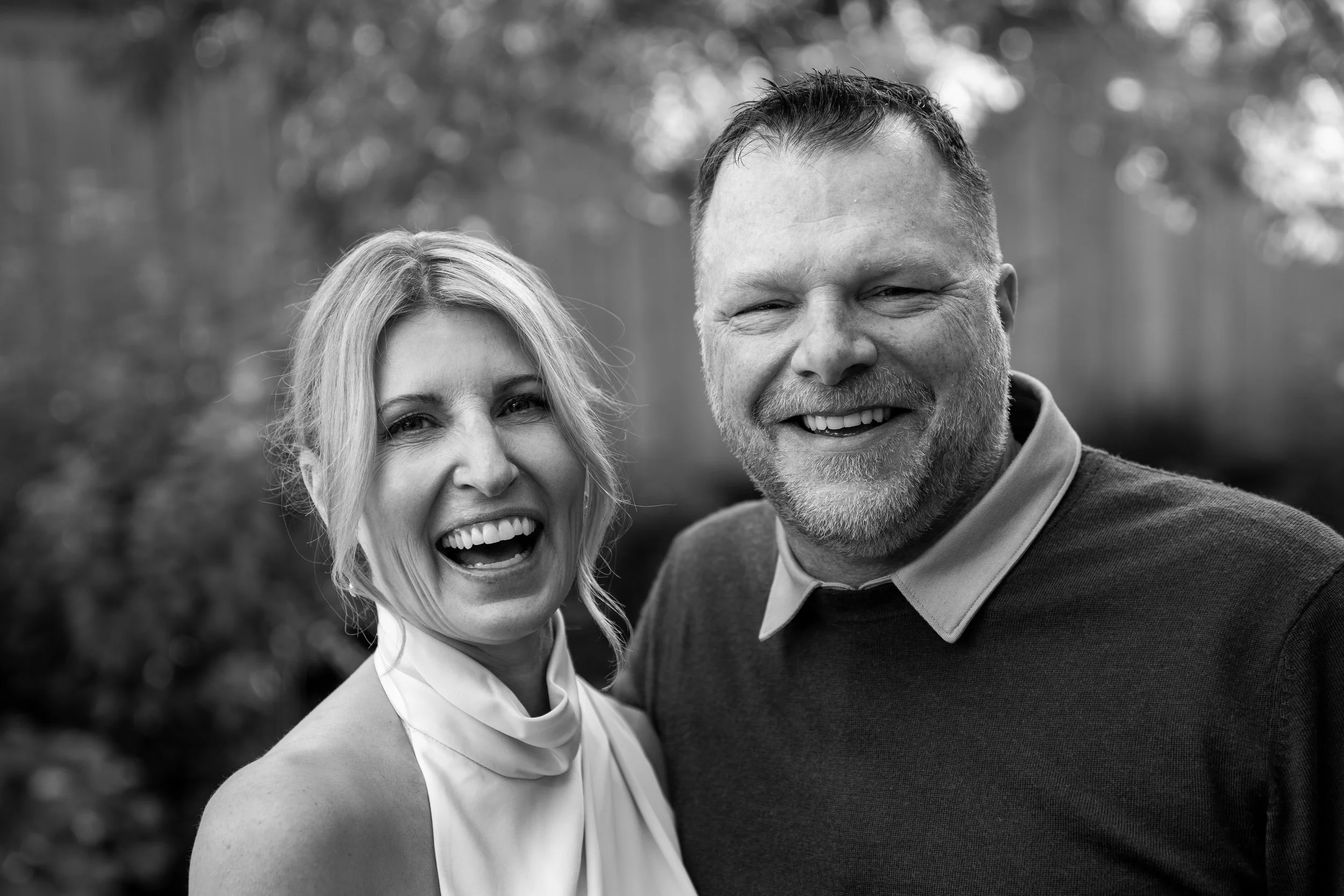 A black and white photo of a smiling woman and man outdoors, close together, both facing the camera.