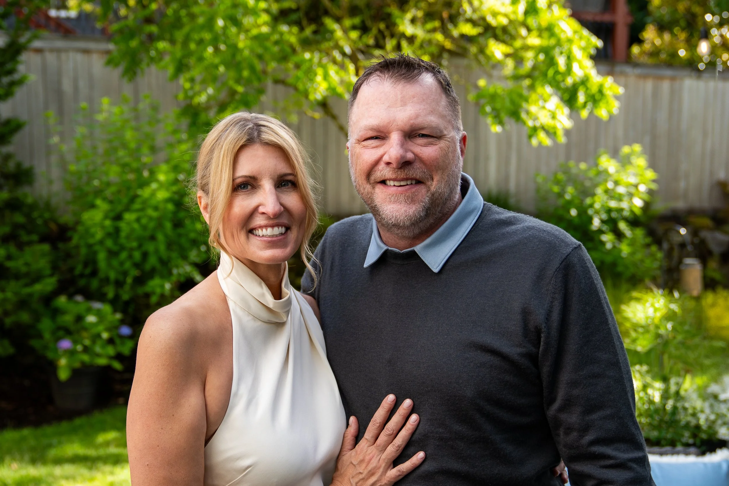 A smiling woman with blonde hair and a man with short hair and a beard, both standing outdoors in a garden with green trees and a wooden fence in the background.