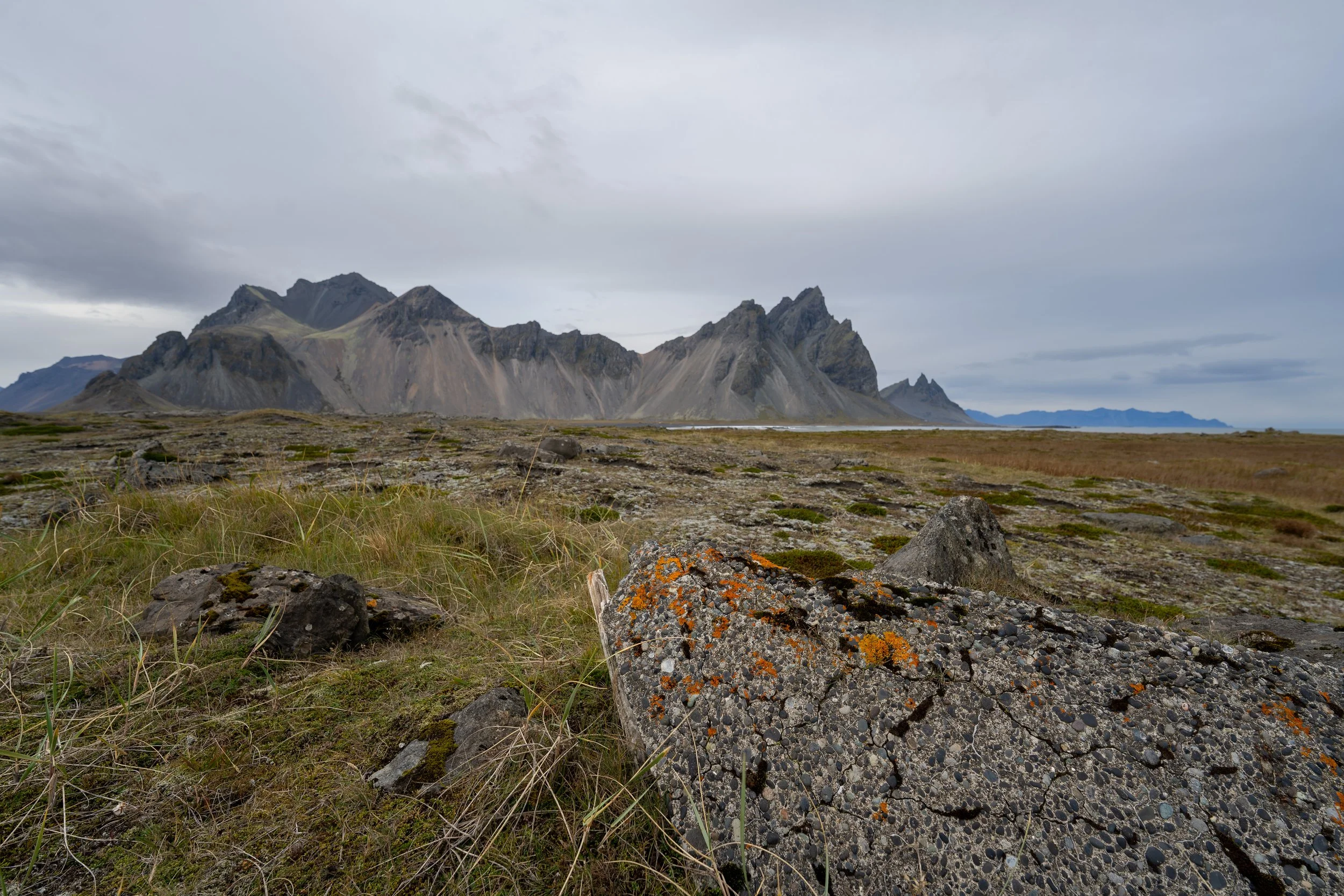A rugged mountain range with jagged peaks and gray slopes on a cloudy day, with a foreground of grass, rocks, and lichen-covered stones.