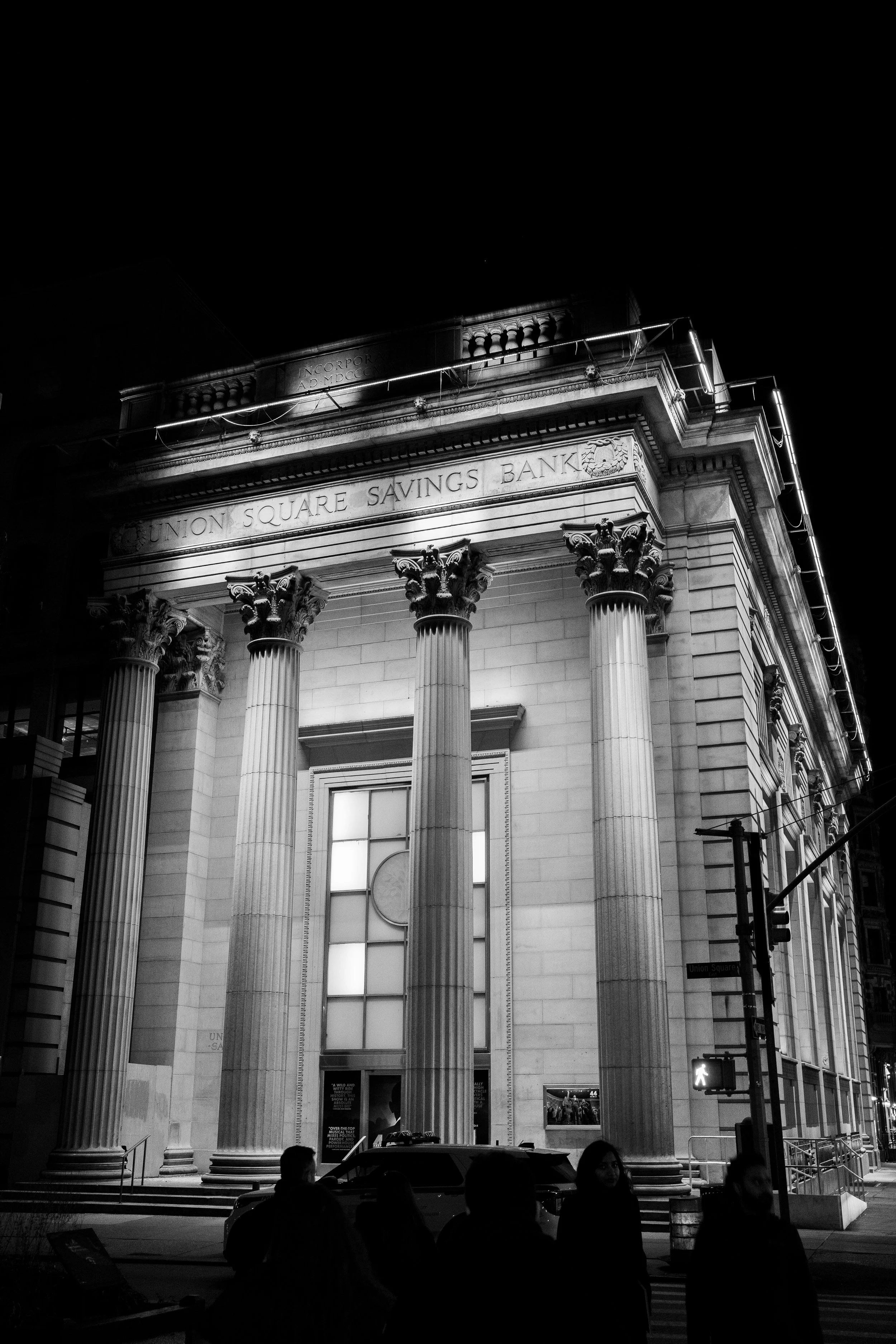 Night view of the Union Square Savings Bank building with classical architectural columns illuminated at night, street pedestrians, and a lit crosswalk signal.