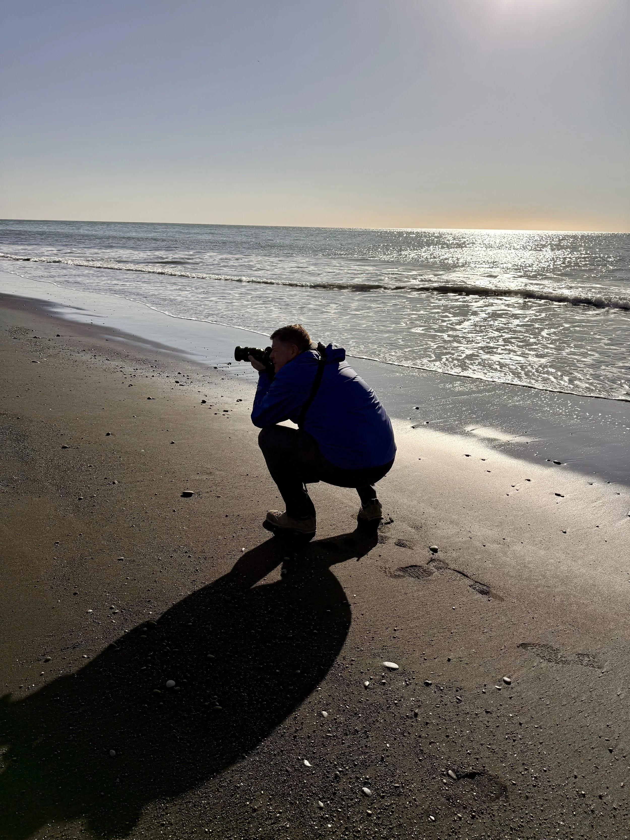 A person crouching on a sandy beach taking a photograph of the ocean at sunset or sunrise, with the sun reflecting off the water and casting a shadow of the person on the sand.