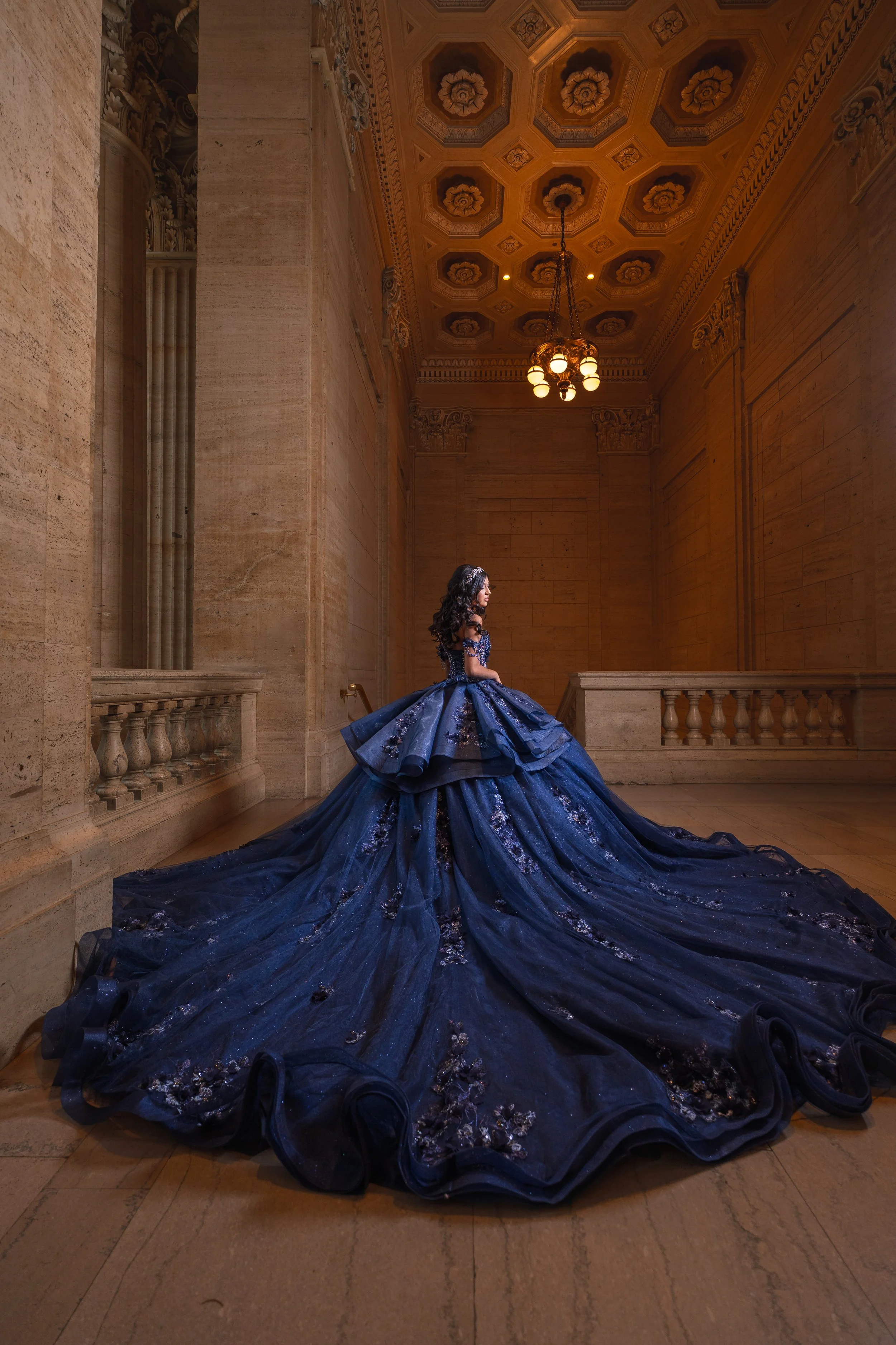 A woman in a dark blue, elaborate ball gown with floral embroidery standing in a grand, ornate hall with high ceilings, chandeliers, and beige stone walls.