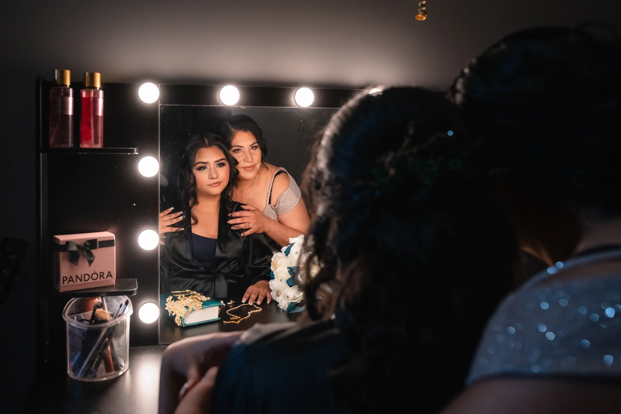 Two women looking into a mirror with lights, one with dark makeup and the other holding her shoulder, surrounded by makeup and jewelry on the vanity.