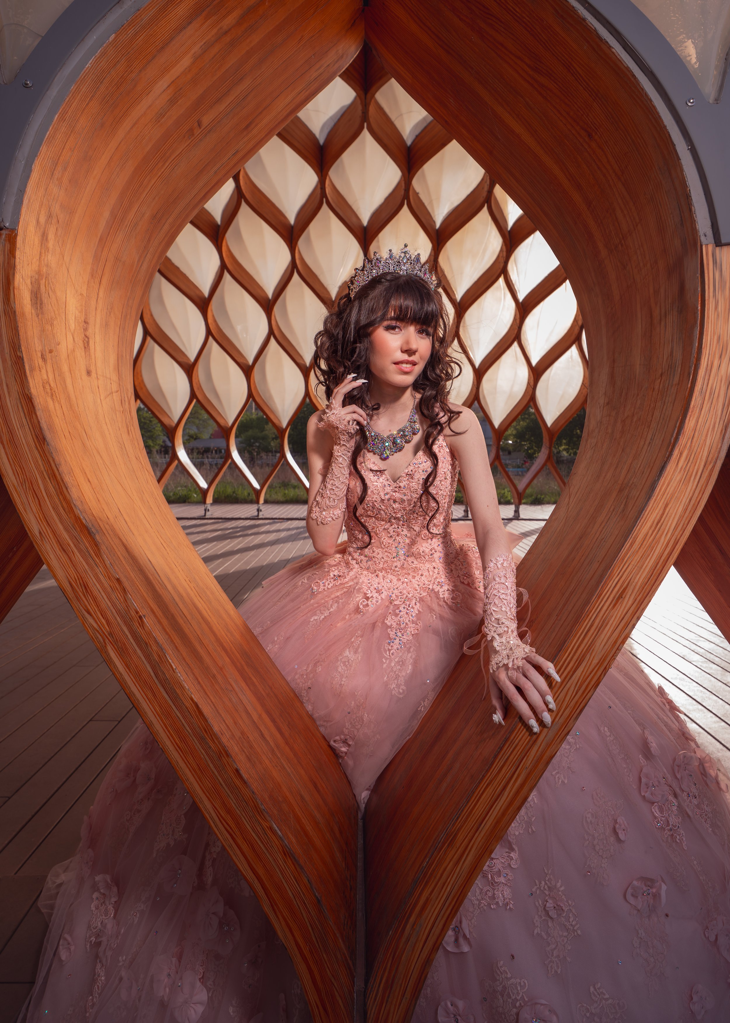Young woman in a pink ball gown with lace and embroidery details, wearing a tiara and a statement necklace, posed inside a wooden heart-shaped structure outdoors.