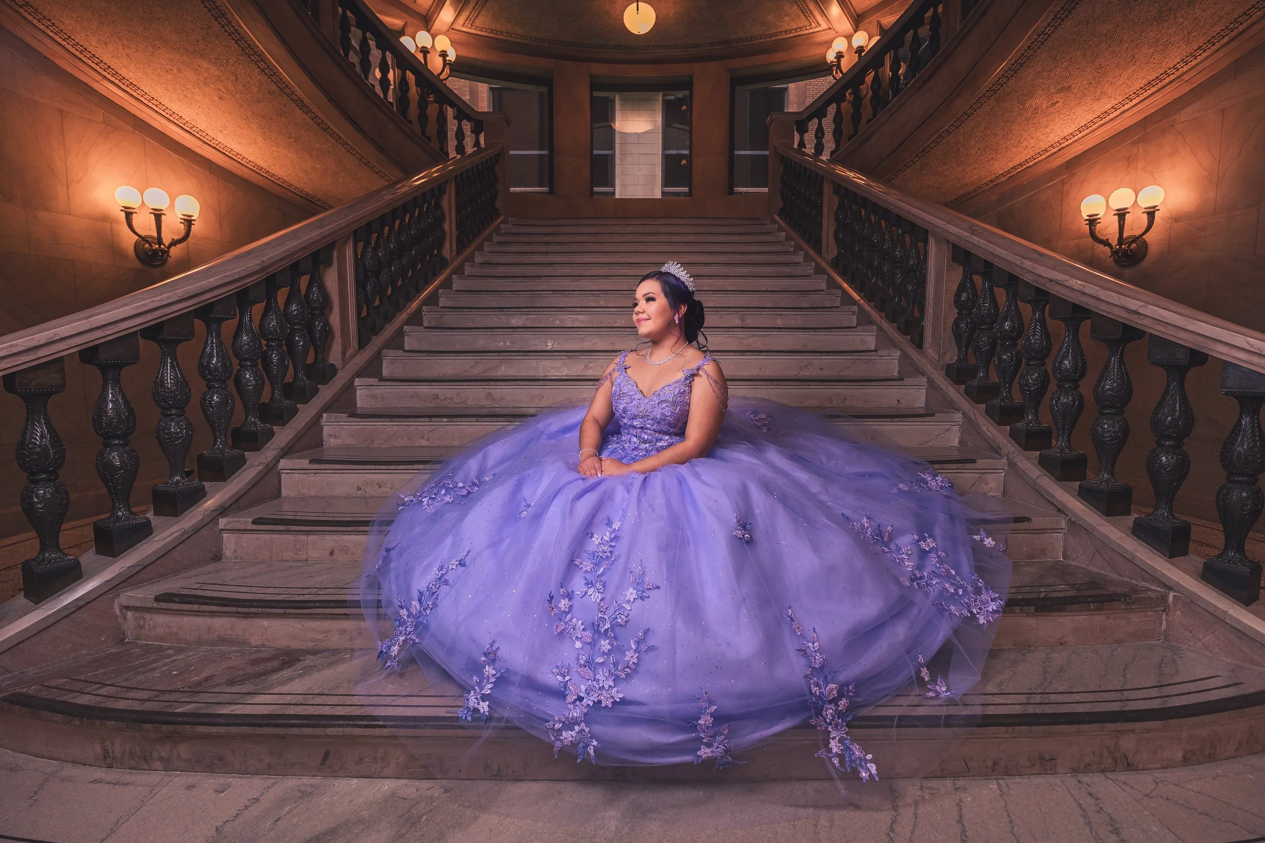 A woman in a purple ball gown and tiara sitting on the stairs of a grand staircase inside a building, with warm lighting and ornate railing.