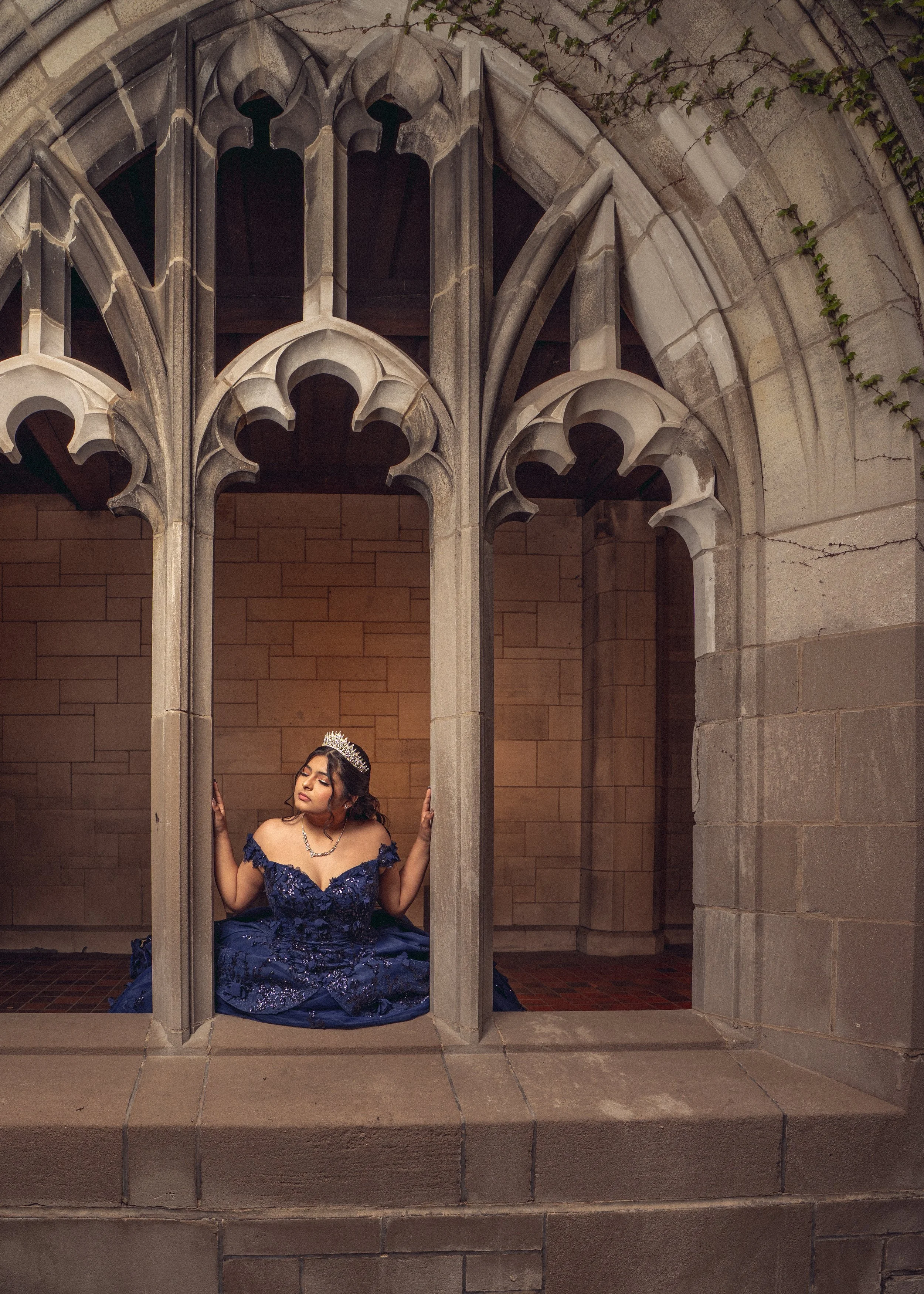 A woman in an elegant navy blue gown and crown poses inside a stone Gothic-style window frame with decorative arches, set against a brick wall and weathered stone exterior with some green vines.