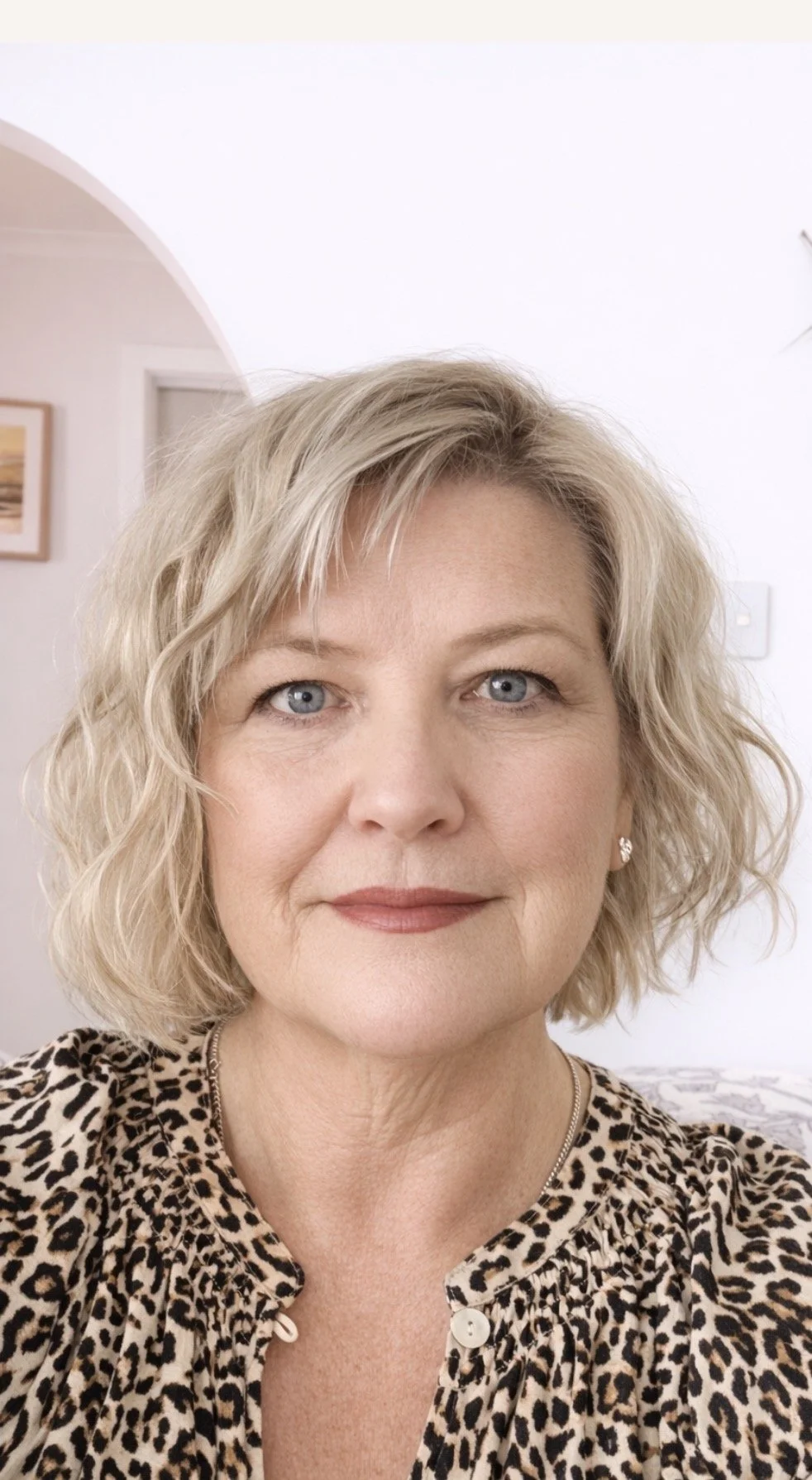 Close-up of a woman with short blonde curly hair, blue eyes, and wearing a leopard print top, standing indoors.