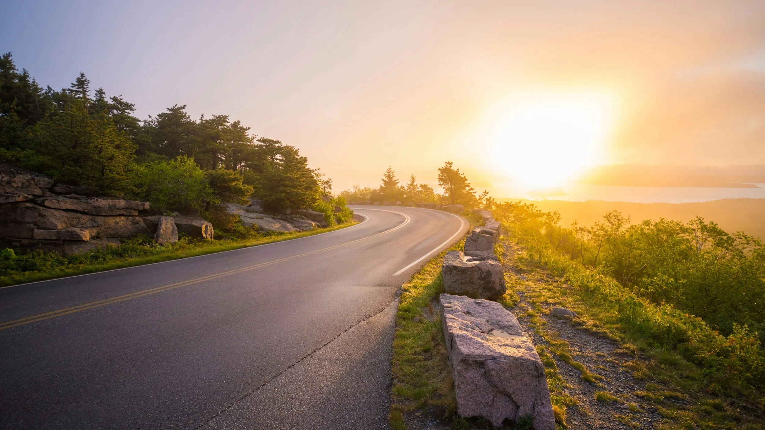 A scenic winding mountain road at sunset with trees along the side and rocky barriers, overlooking a landscape with a body of water and hills in the distance.