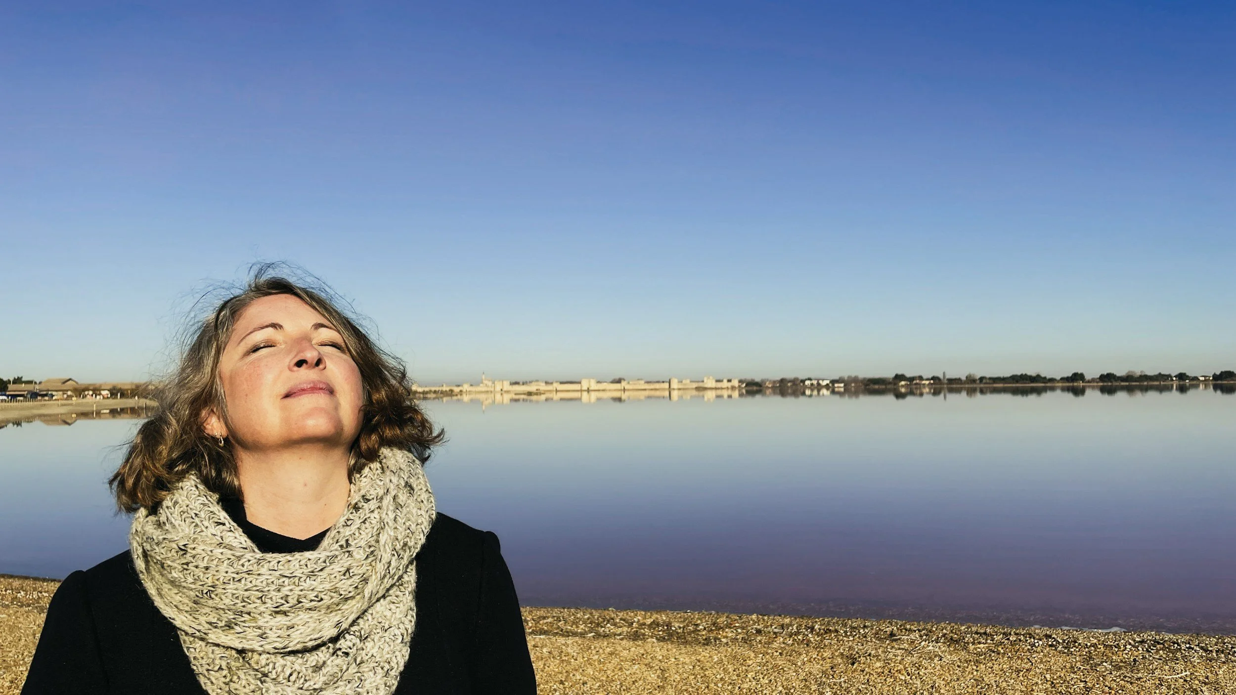 A woman with curly hair and closed eyes, smiling peacefully, standing near a body of water on a sunny day, wearing a knitted scarf and a black top.
