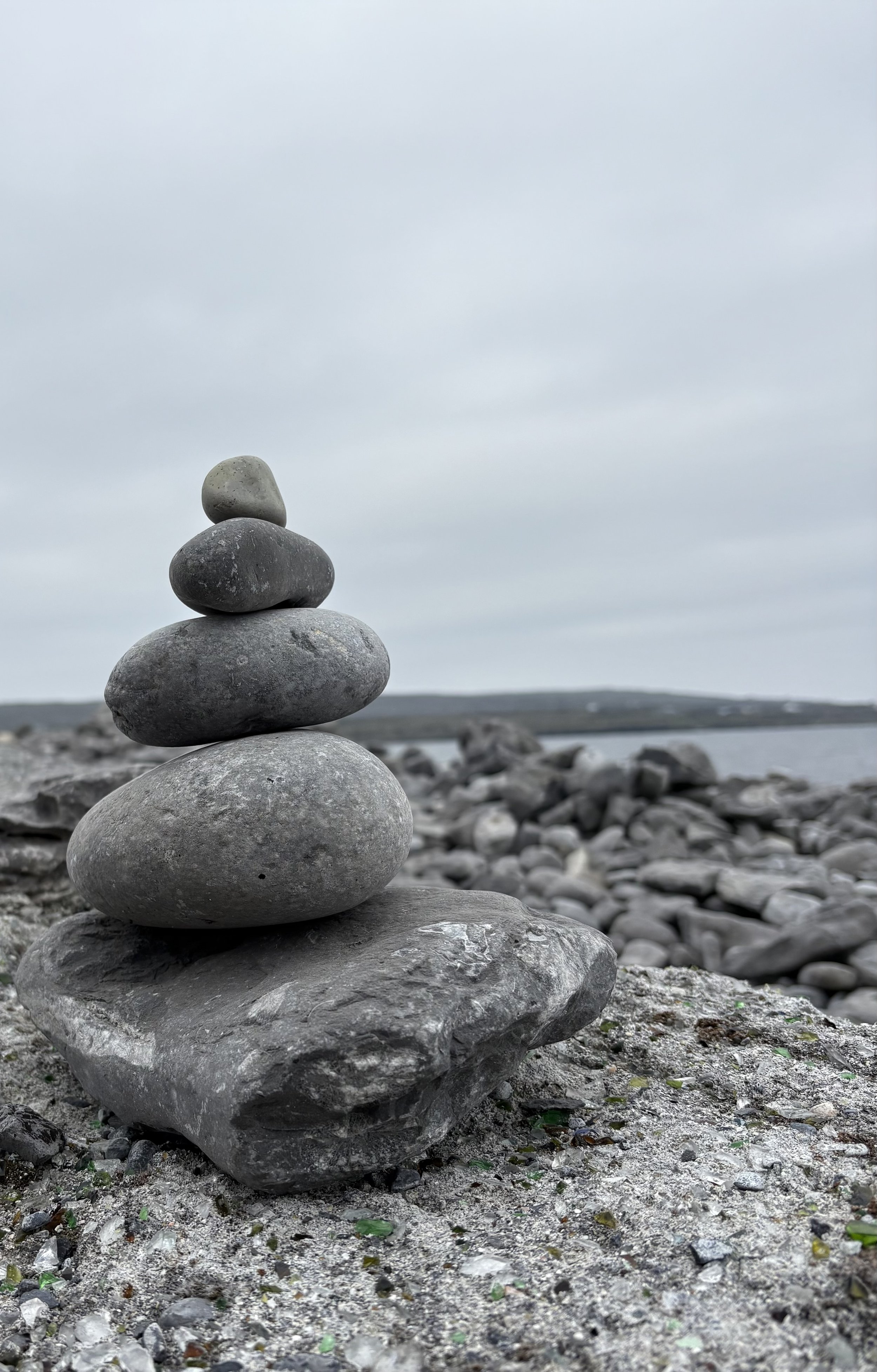 A balanced stack of six smooth gray stones arranged vertically on a rocky beach, with a gray overcast sky and distant shoreline in the background.