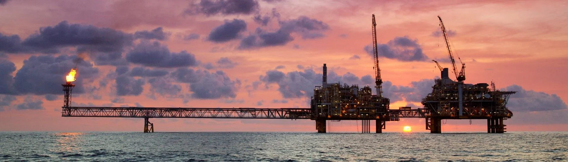 Offshore oil rig during sunset with drilling platform, cranes, and a burning flame from a flare stack, set against cloudy sky over ocean.