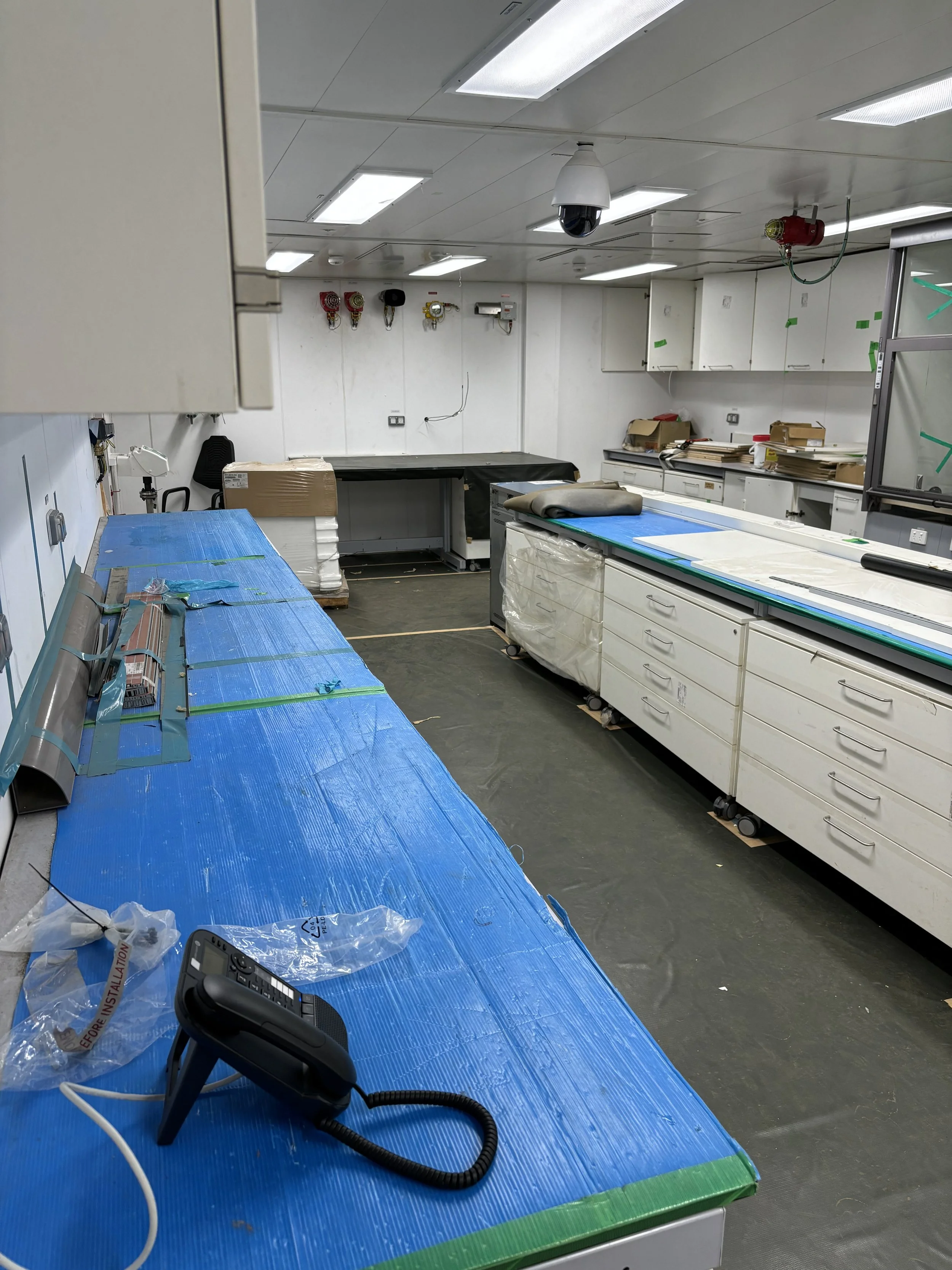 A clean, organized laboratory or medical workspace with various equipment, cabinets, and work surfaces. There is a blue-covered table with small parts, a cordless phone, and an electronic device in the foreground.