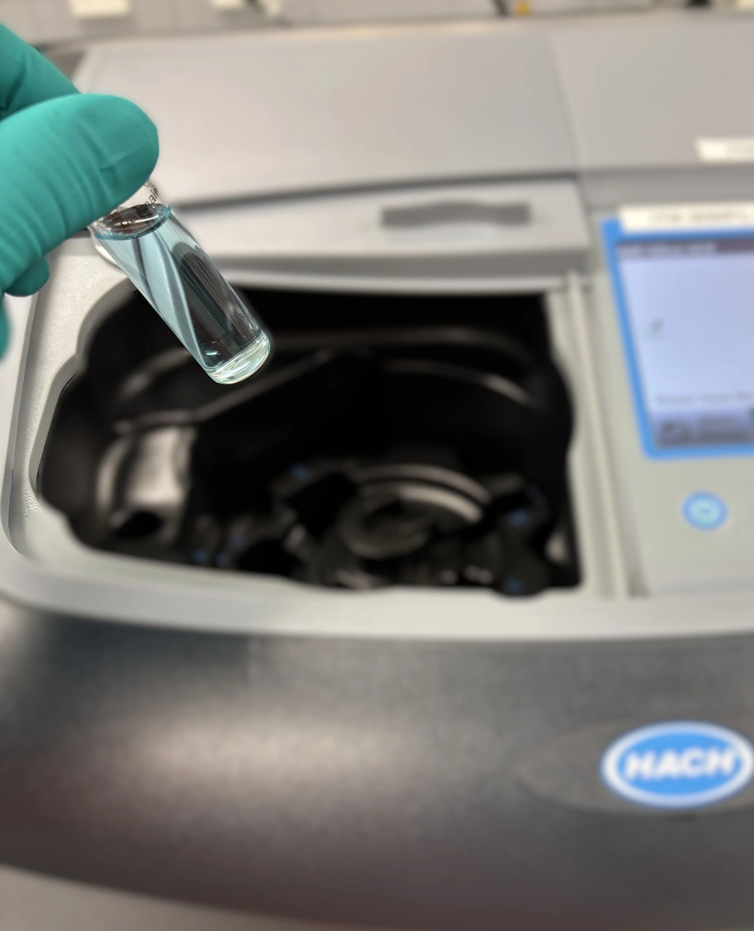 Close-up of a gloved hand holding a small glass vial with a clear liquid inside, being positioned above a centrifuge machine in a laboratory setting.