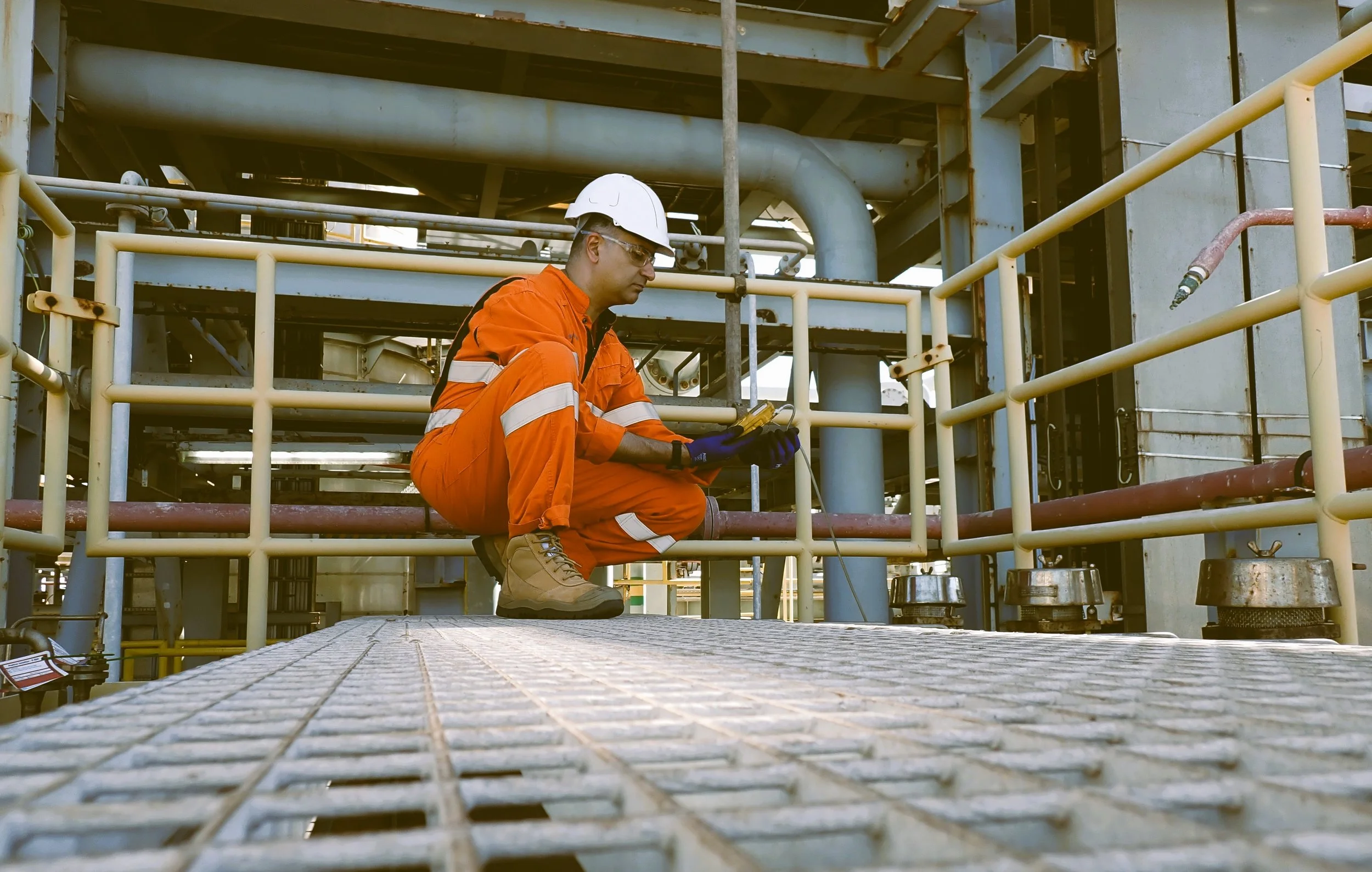 A male industrial worker wearing an orange safety jumpsuit, white hard hat, safety glasses, and gloves is crouched down on a metal platform inspecting equipment, with a background of industrial pipes and structures.