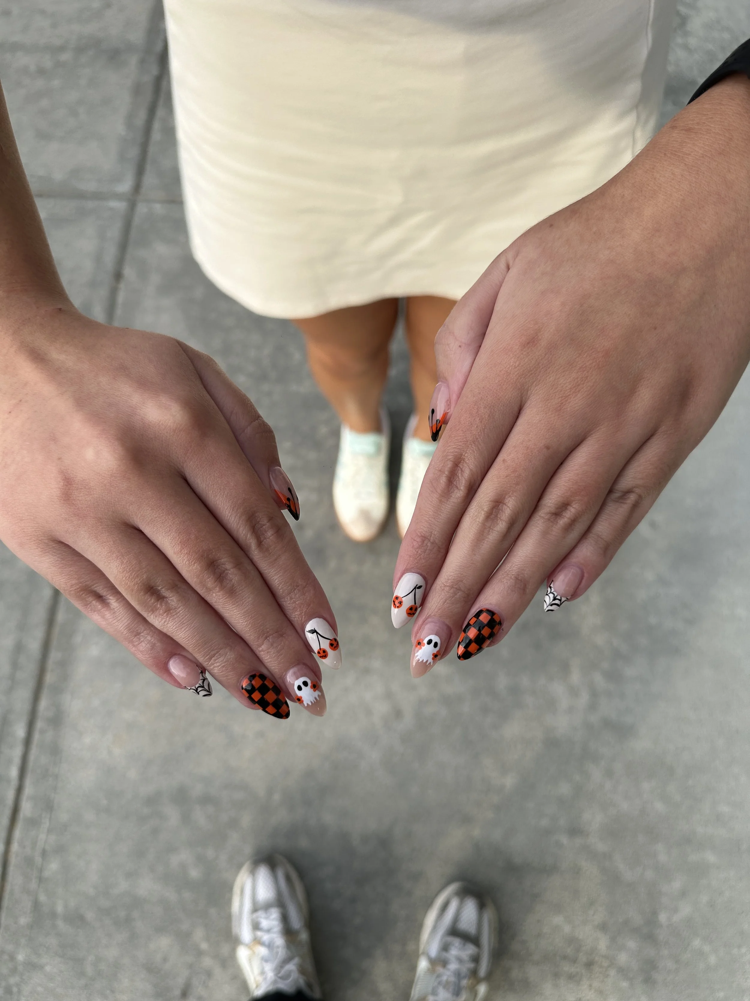 Person showing hands with Halloween-themed nail art, including little ghosts, pumpkins, spiders, and orange-black checkered patterns, while standing on a sidewalk.
