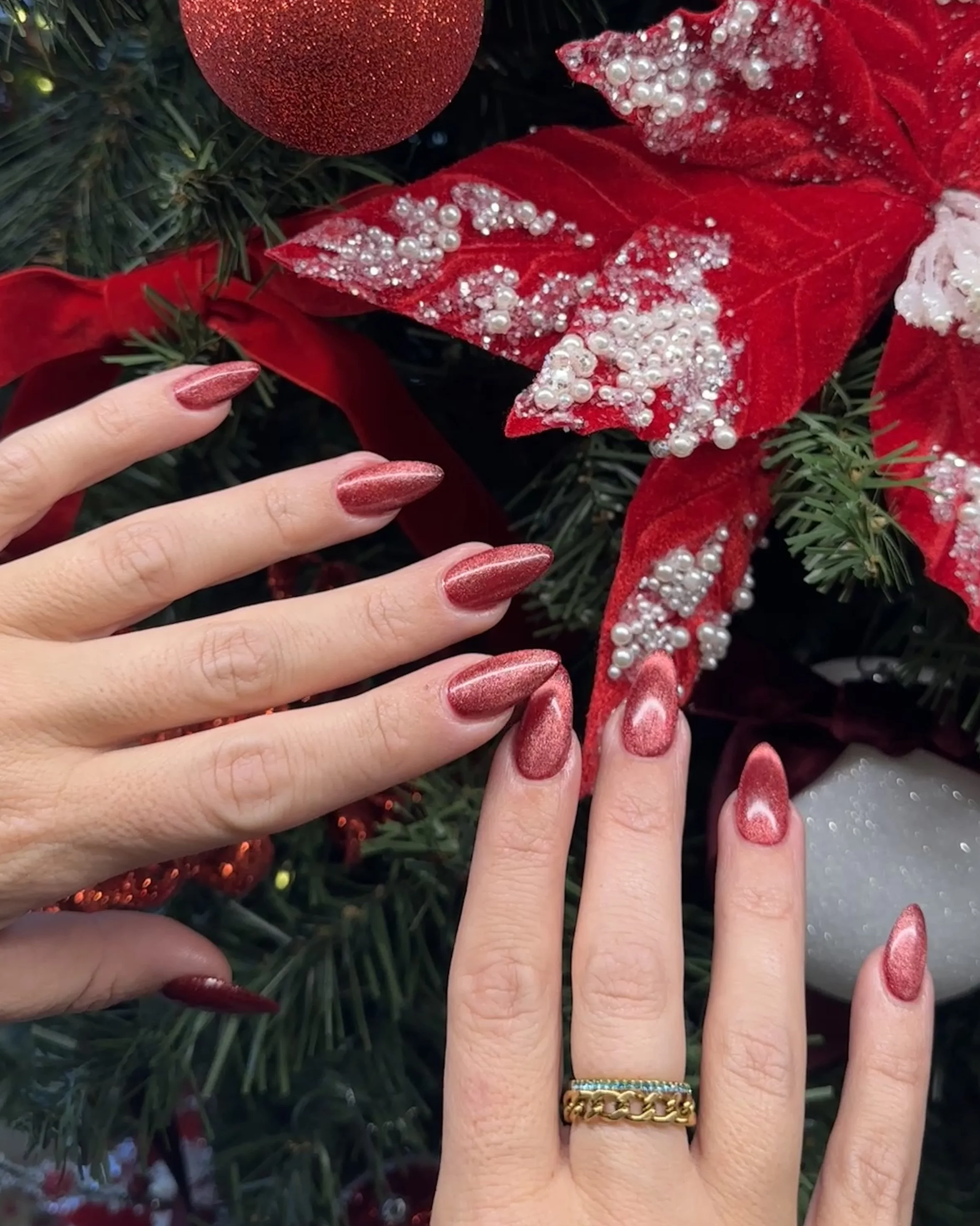 Close-up of two hands with red metallic Christmas-themed nails, touching a red poinsettia flower with pearl embellishments, surrounded by Christmas decorations including a red ornament and a white bauble.