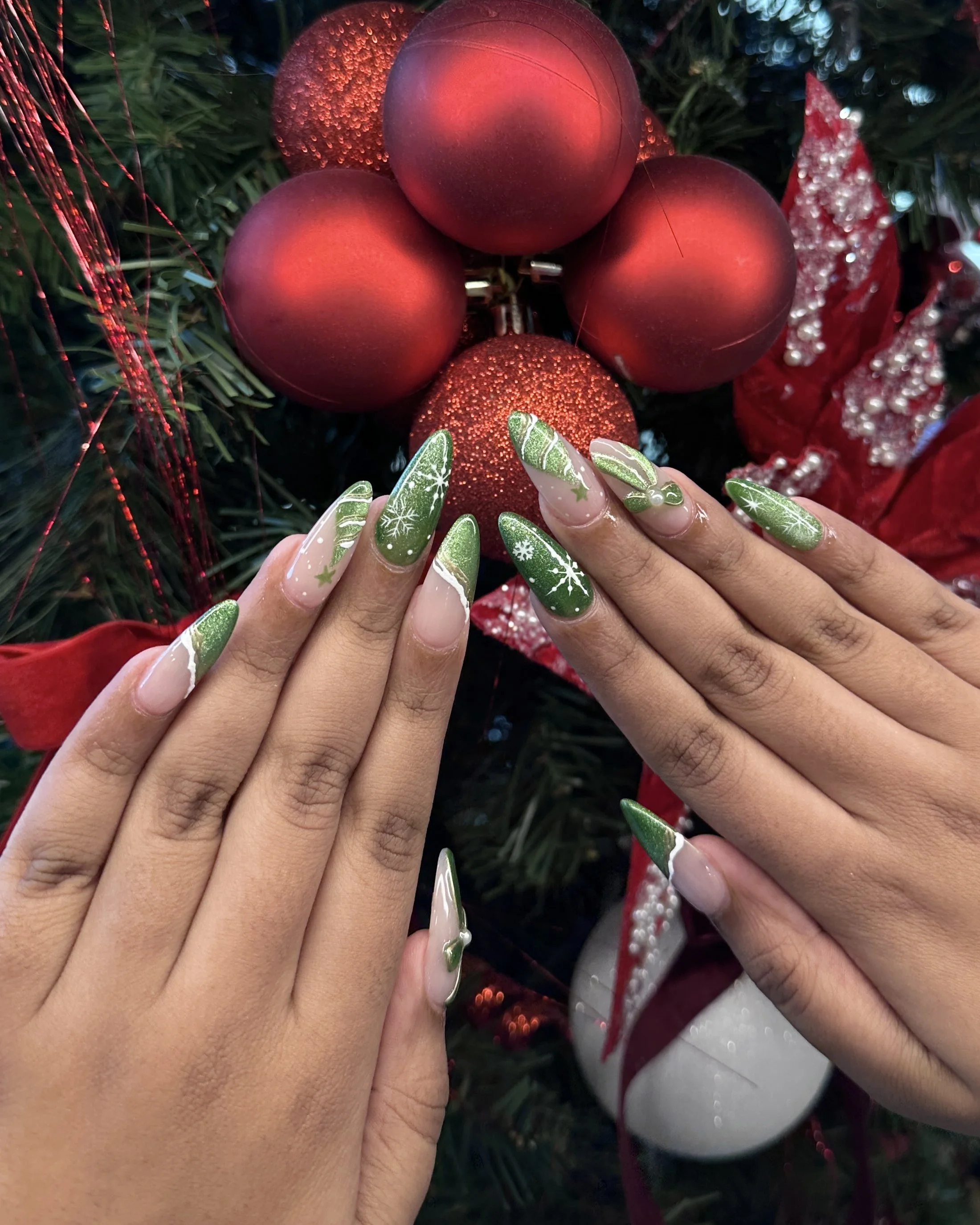 Person showing off festive green and white Christmas nail art with decorated nails in front of a Christmas tree with red ornaments and ribbons.