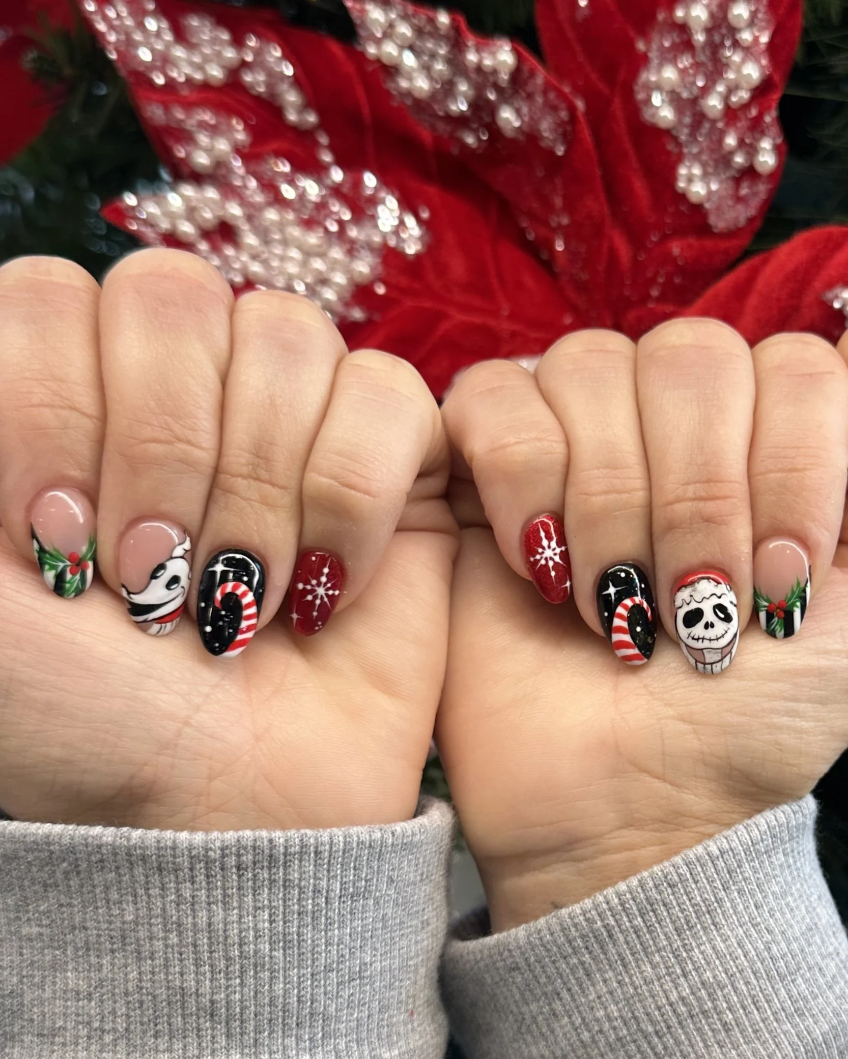Nail art with Christmas-themed designs including holly, a reindeer, candy canes, snowflakes, a skull with Santa hat, and holiday patterns, against a background of a red poinsettia flower.