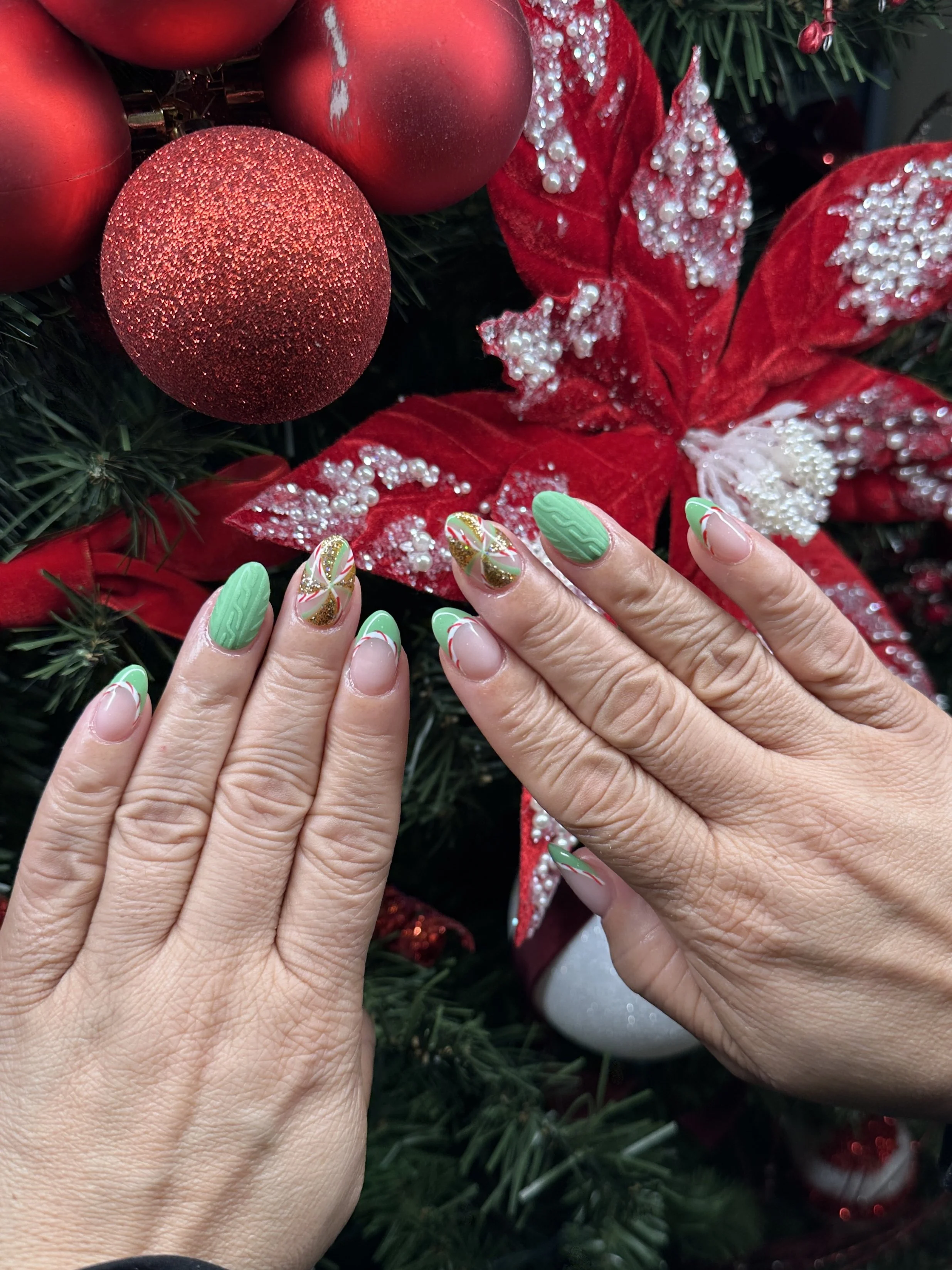 Hands with decorated nails in front of Christmas decorations including red ornaments and poinsettia flower.