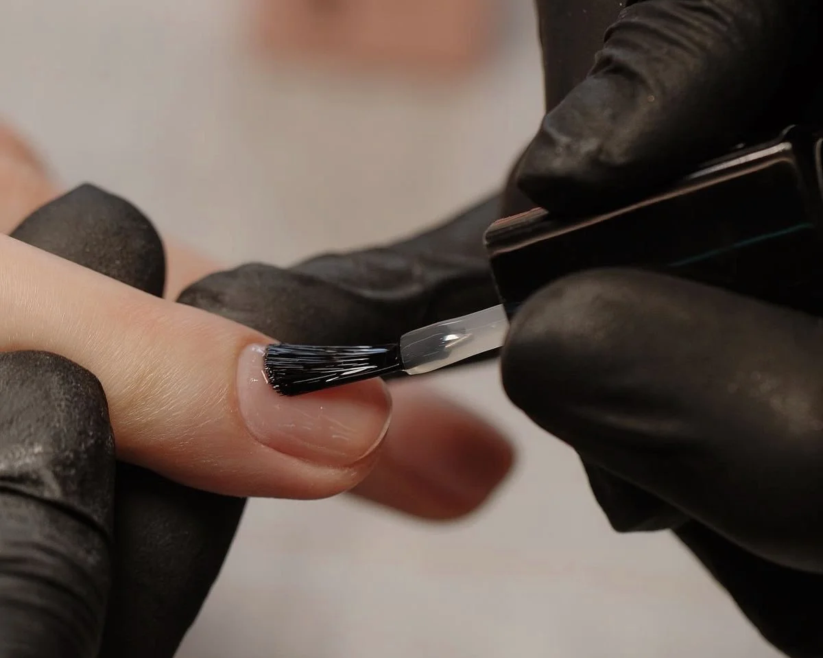 Close-up of a person applying black gel nail polish with a brush to a fingernail, wearing black gloves.