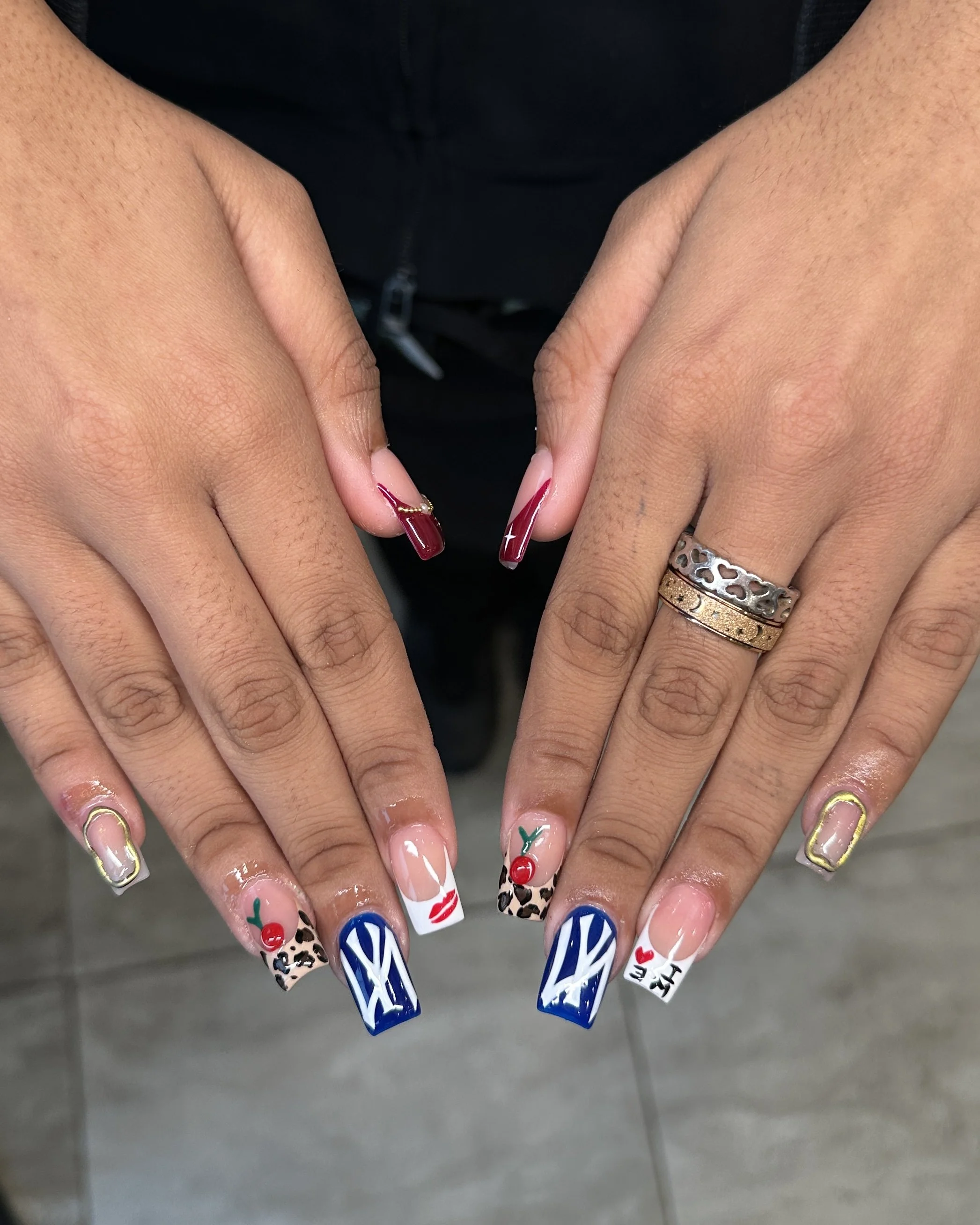 Close-up of hands showing decorated acrylic nails with various designs including cherries, animal print, blue pattern, lips, and 'LOVE' text, and rings on the fingers.