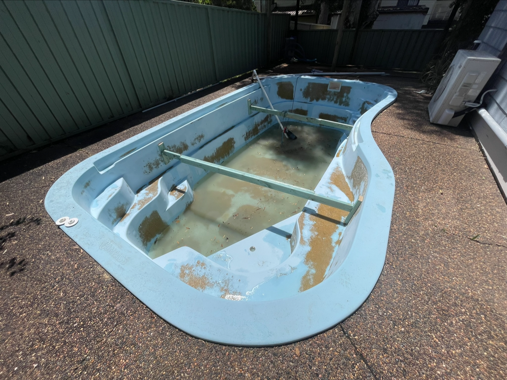 Old, empty hot tub with dirty water, algae, and debris inside, situated on a concrete surface outdoors, surrounded by a green fence and outdoor equipment.