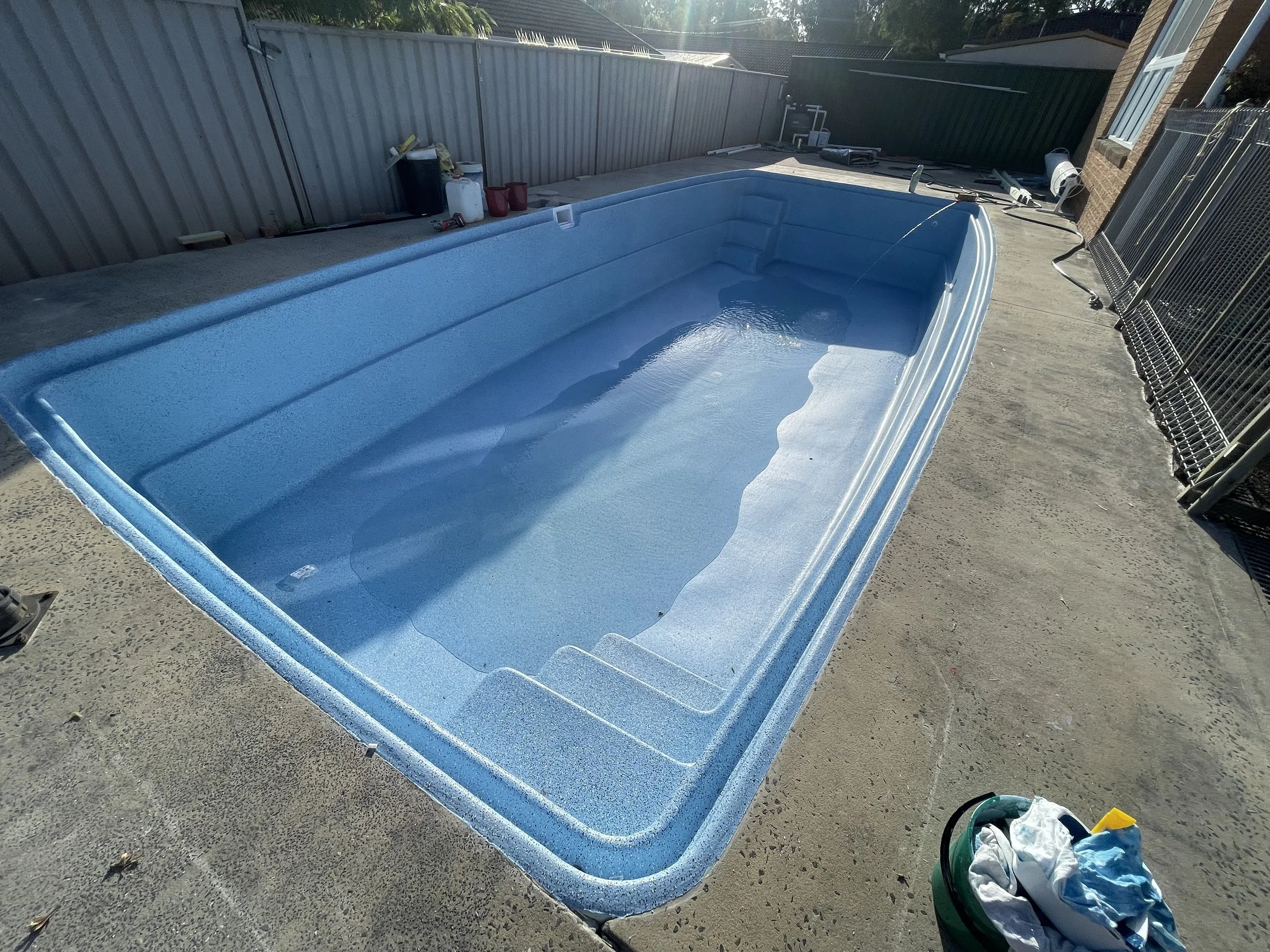 An empty blue swimming pool installed outdoors on a concrete deck, surrounded by a metal fence and some tools and supplies nearby.