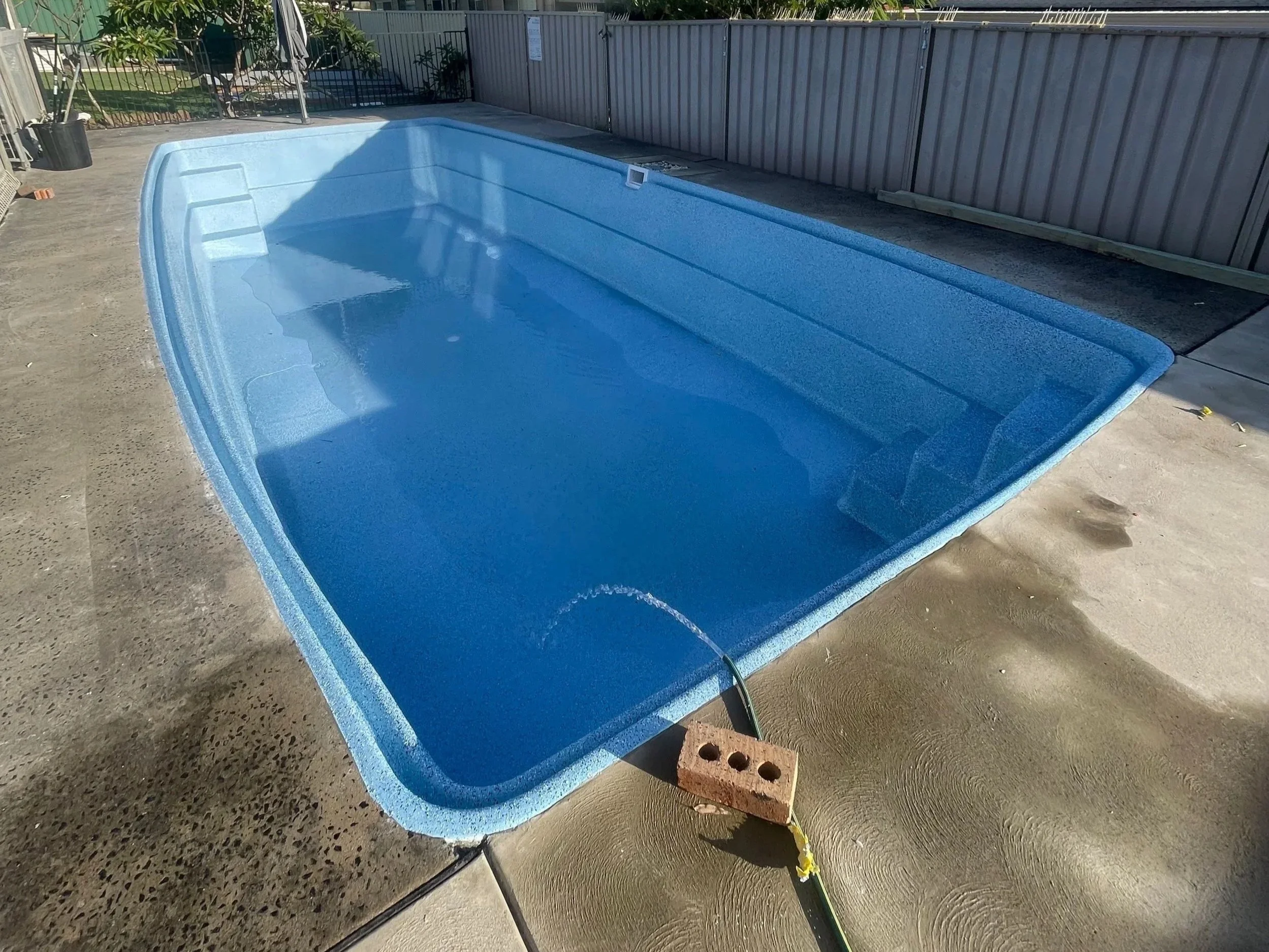 Empty rectangular blue swimming pool on concrete with brick and hose nearby, surrounded by metal fence and some trees.