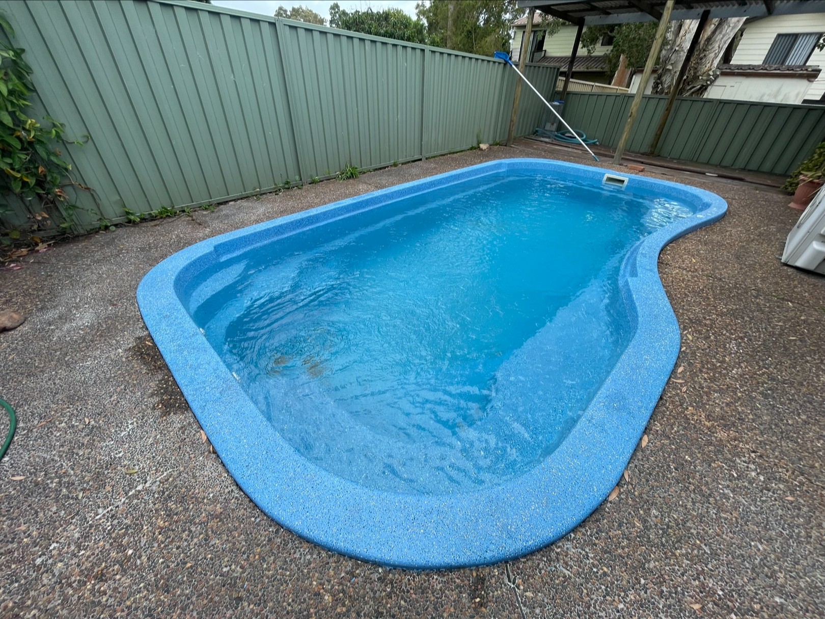 Blue in-ground swimming pool in a backyard, surrounded by a pebble-textured concrete deck, with a green fence, some plants, and a house visible in the background.