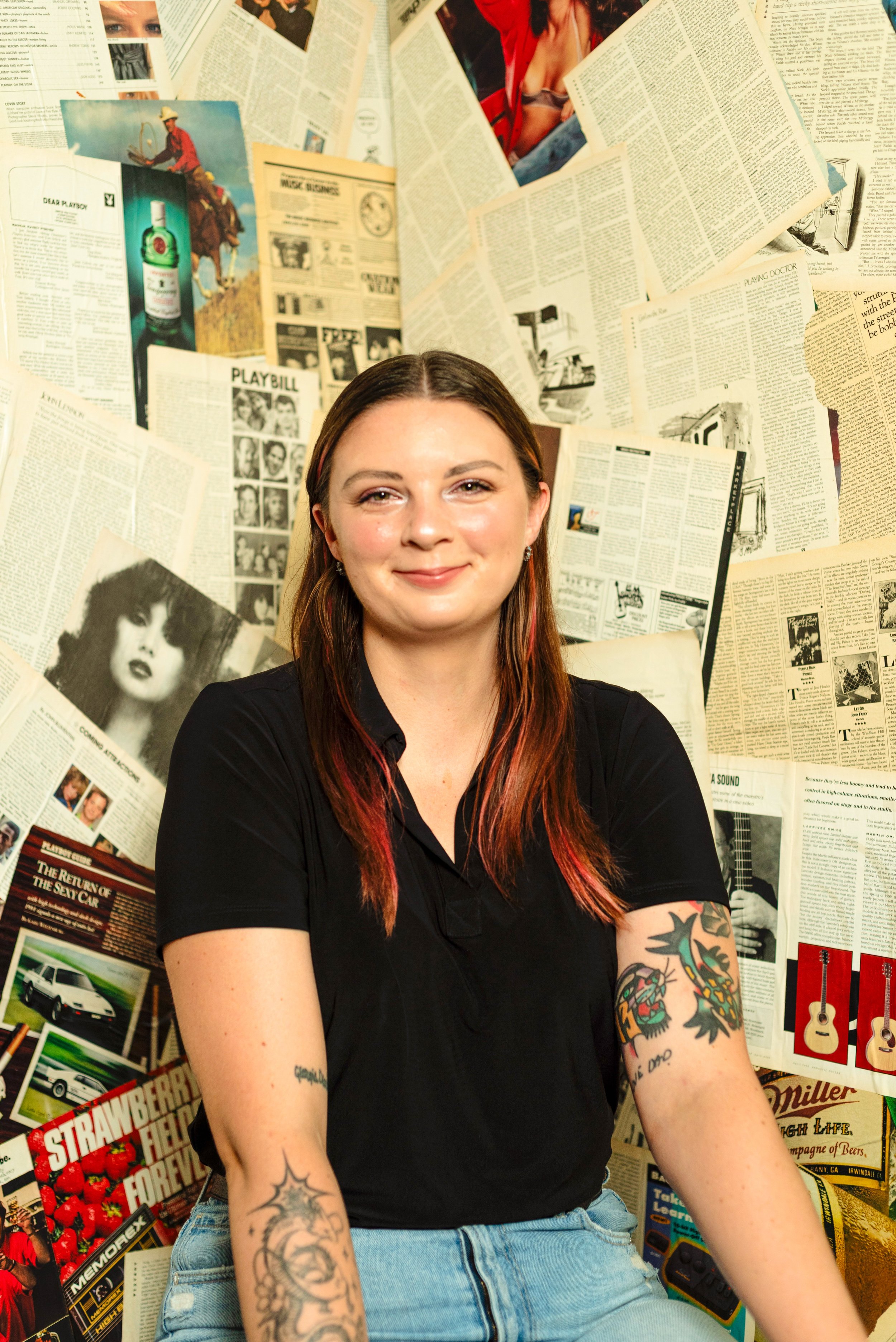 A woman with brown hair and tattoos on her arms, wearing a black shirt and blue jeans, sitting in front of a background made of newspapers and magazine pages.