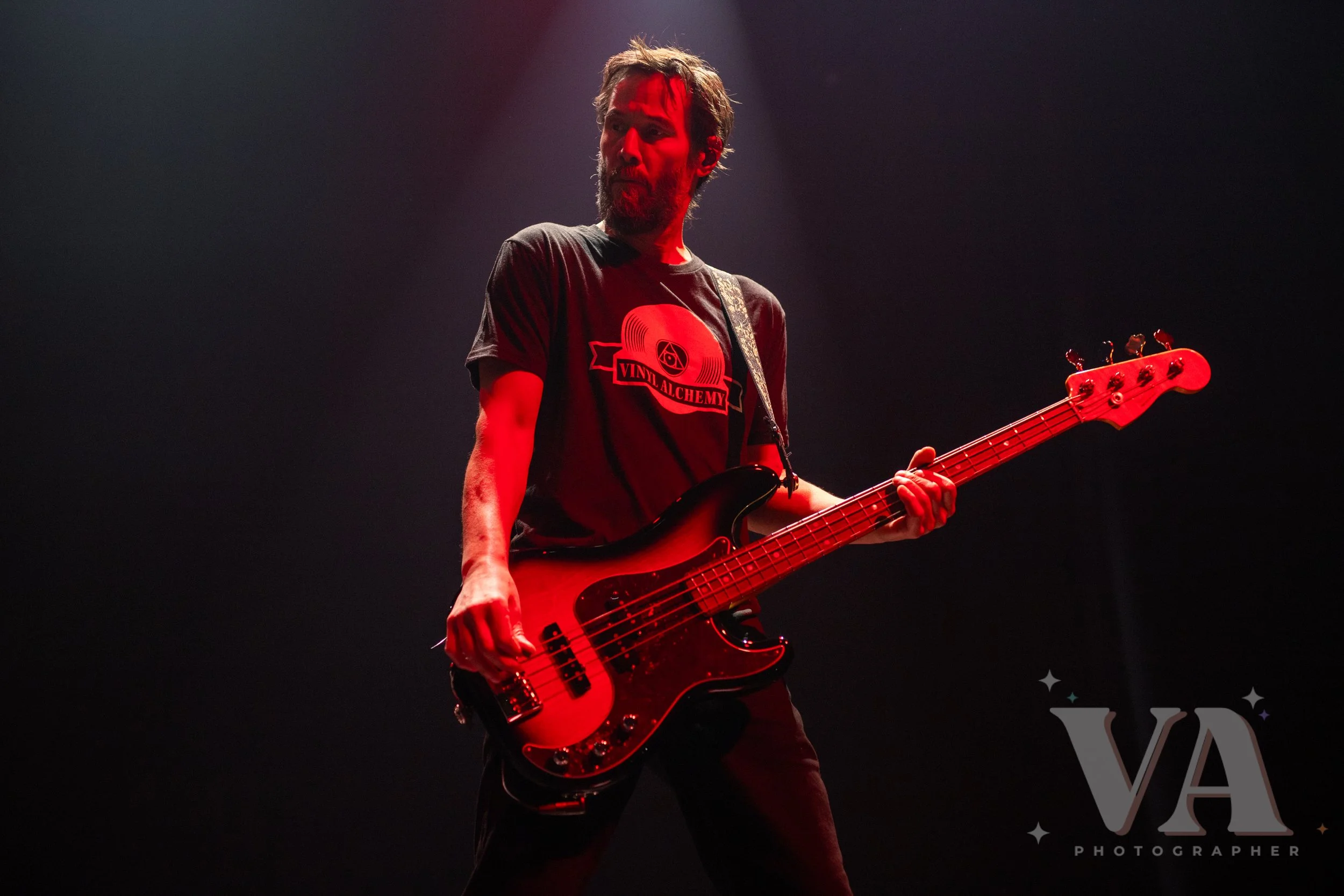 A man playing a bass guitar on stage with red lighting and dark background.