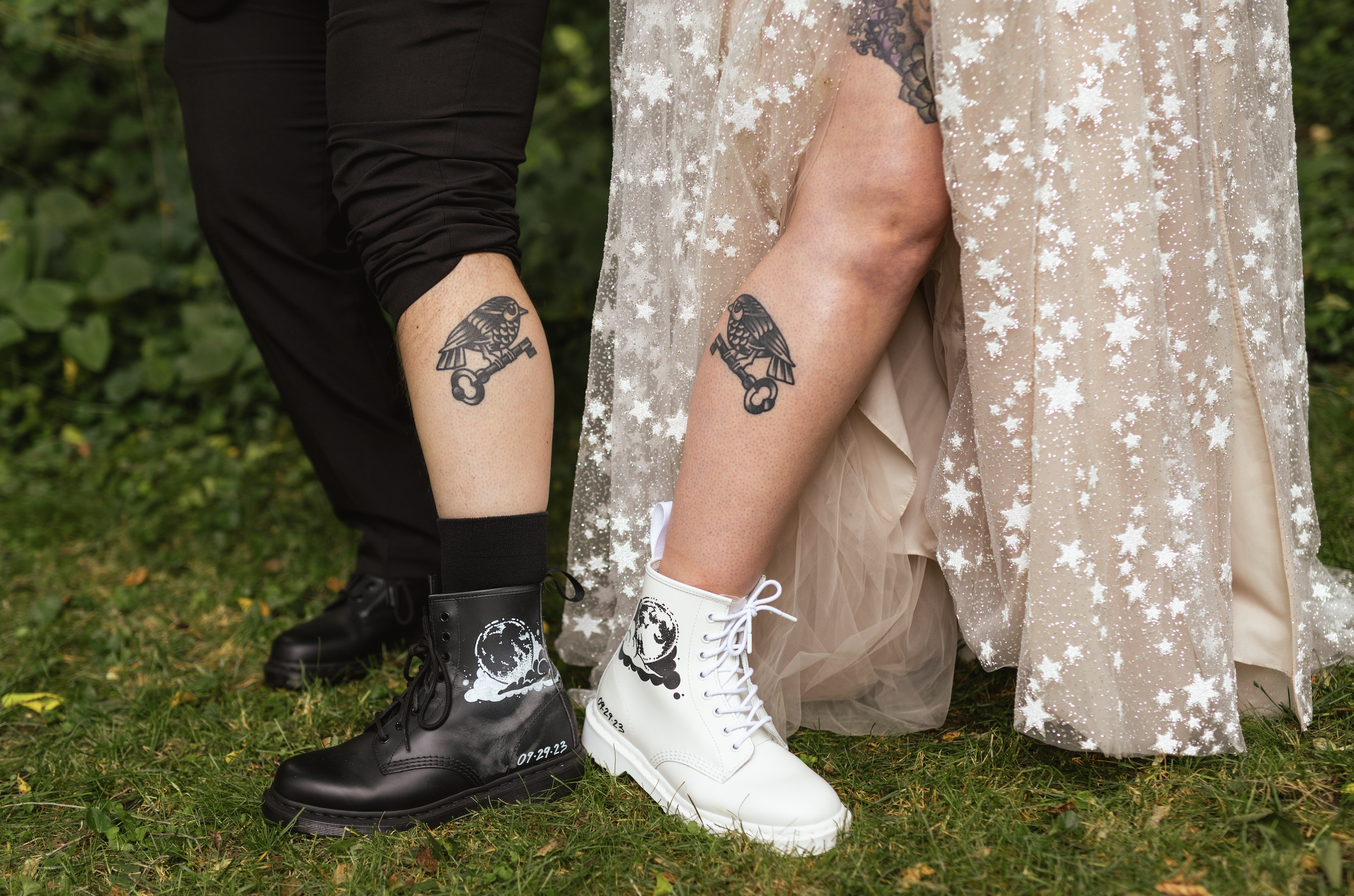 Close-up of two people showing their tattoos of birds holding keys on their legs, wearing black and white combat boots with celestial designs, outdoors on grassy ground.