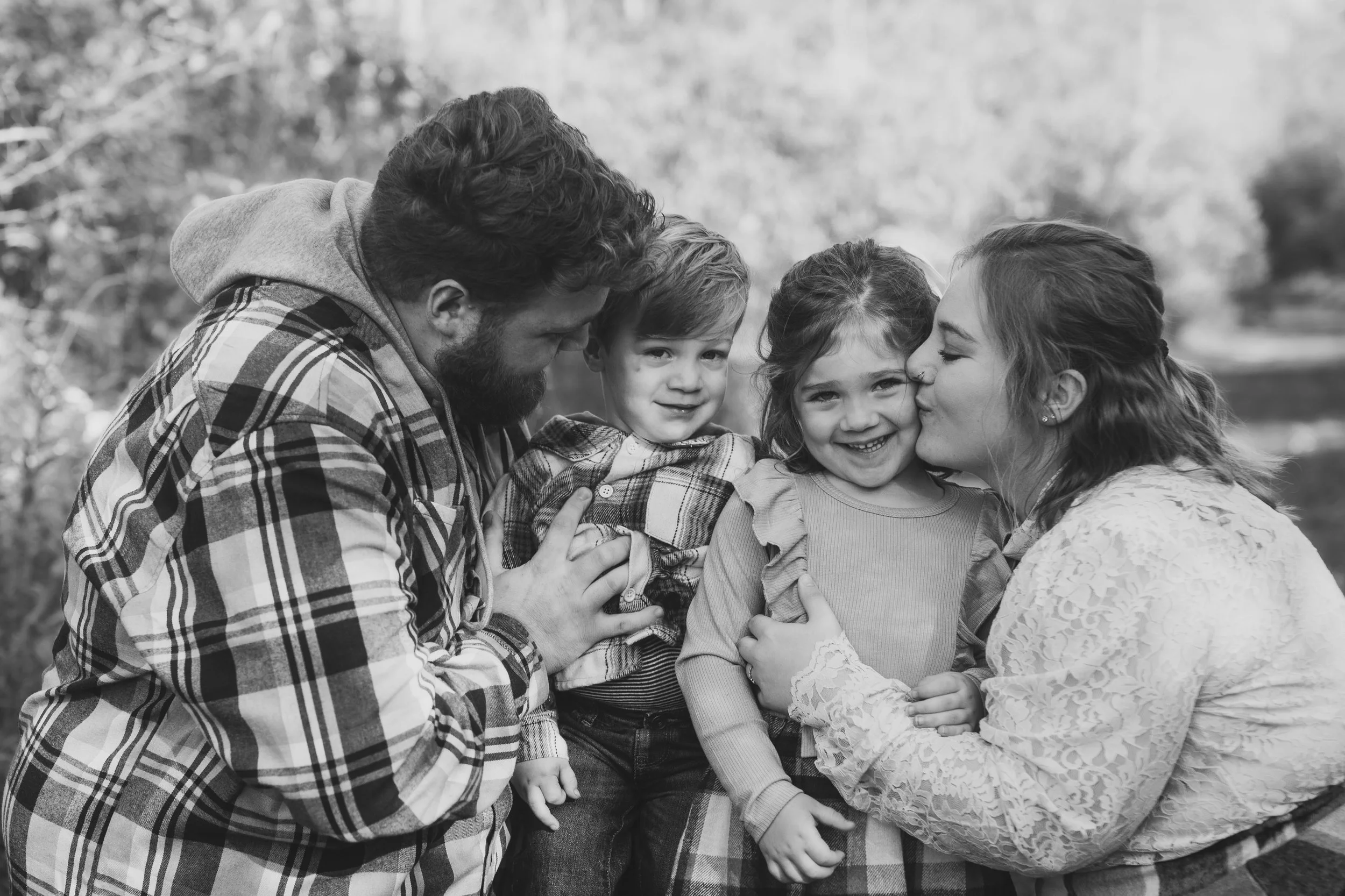 Family of four outdoors, parents with two young children, woman kissing girl, man smiling, all in casual clothing.