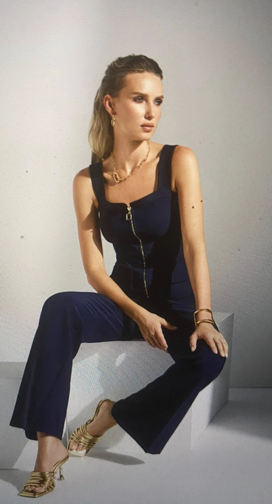 A woman with blonde hair sitting on a low white platform in a black sleeveless jumpsuit with a front zipper and gold accessories, including heels, a necklace, bracelets, and earrings, against a plain gray background.