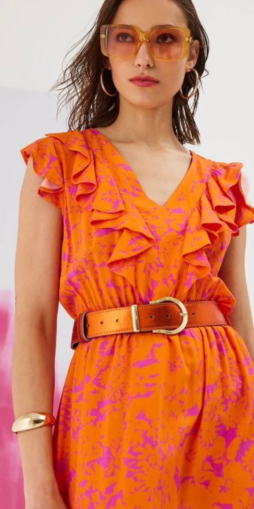 A woman wearing a bright orange dress with purple abstract patterns, ruffled sleeves, and a brown belt, accessorized with large yellow sunglasses, hoop earrings, and a gold bracelet.