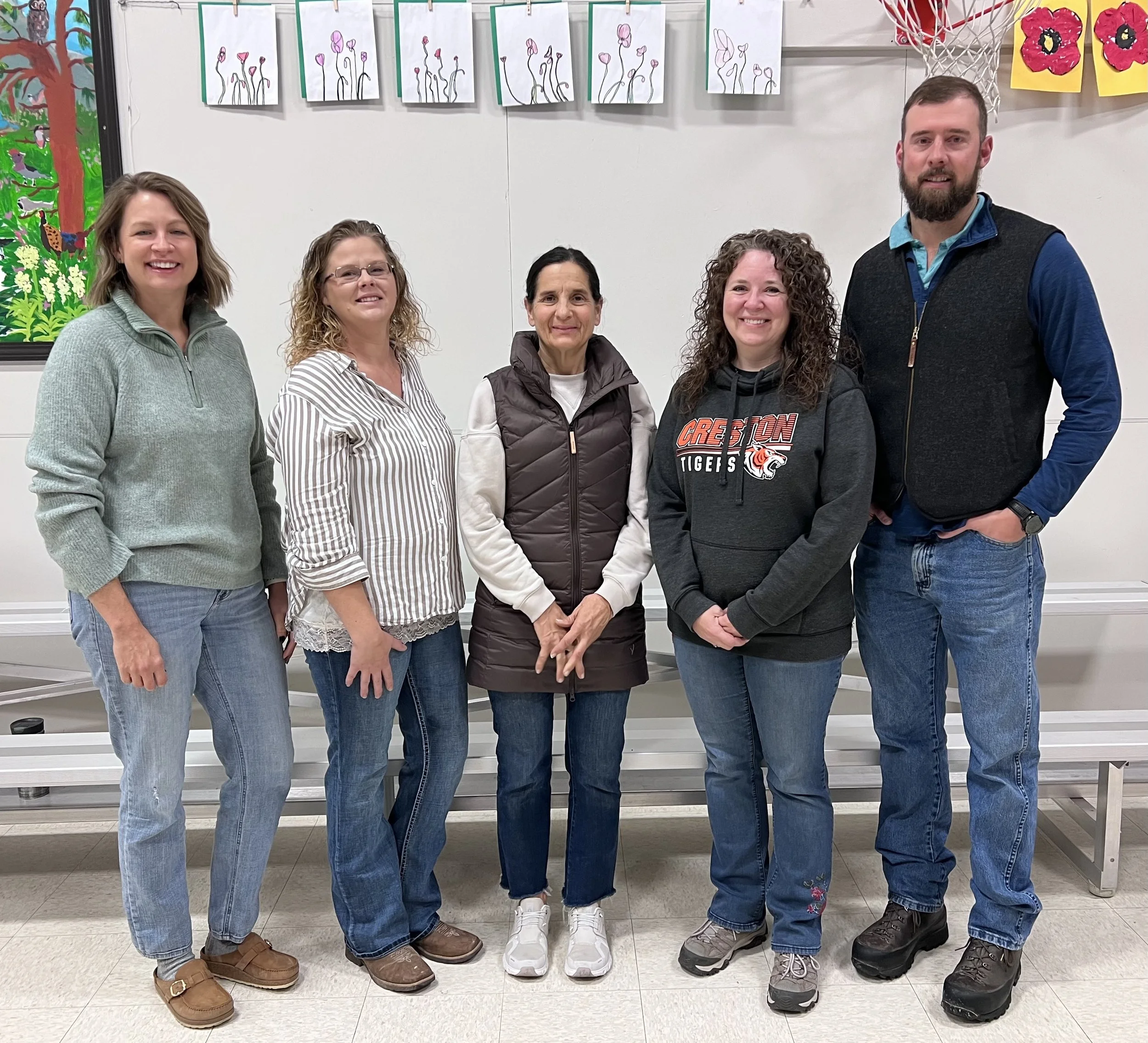 A group of five people standing indoors in front of a wall with children's artwork. They are smiling and dressed casually.