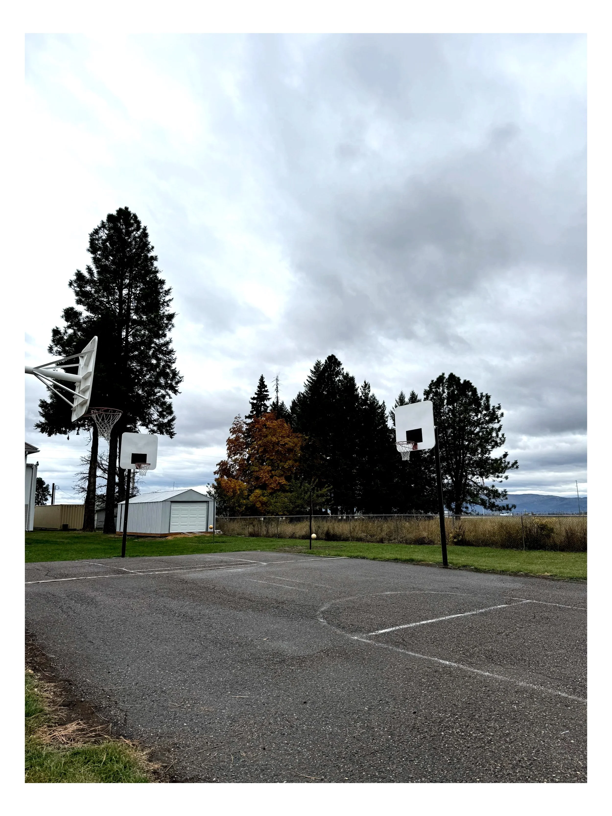 Two empty basketball courts with hoops and backboards in a parking lot, surrounded by trees under a cloudy sky.