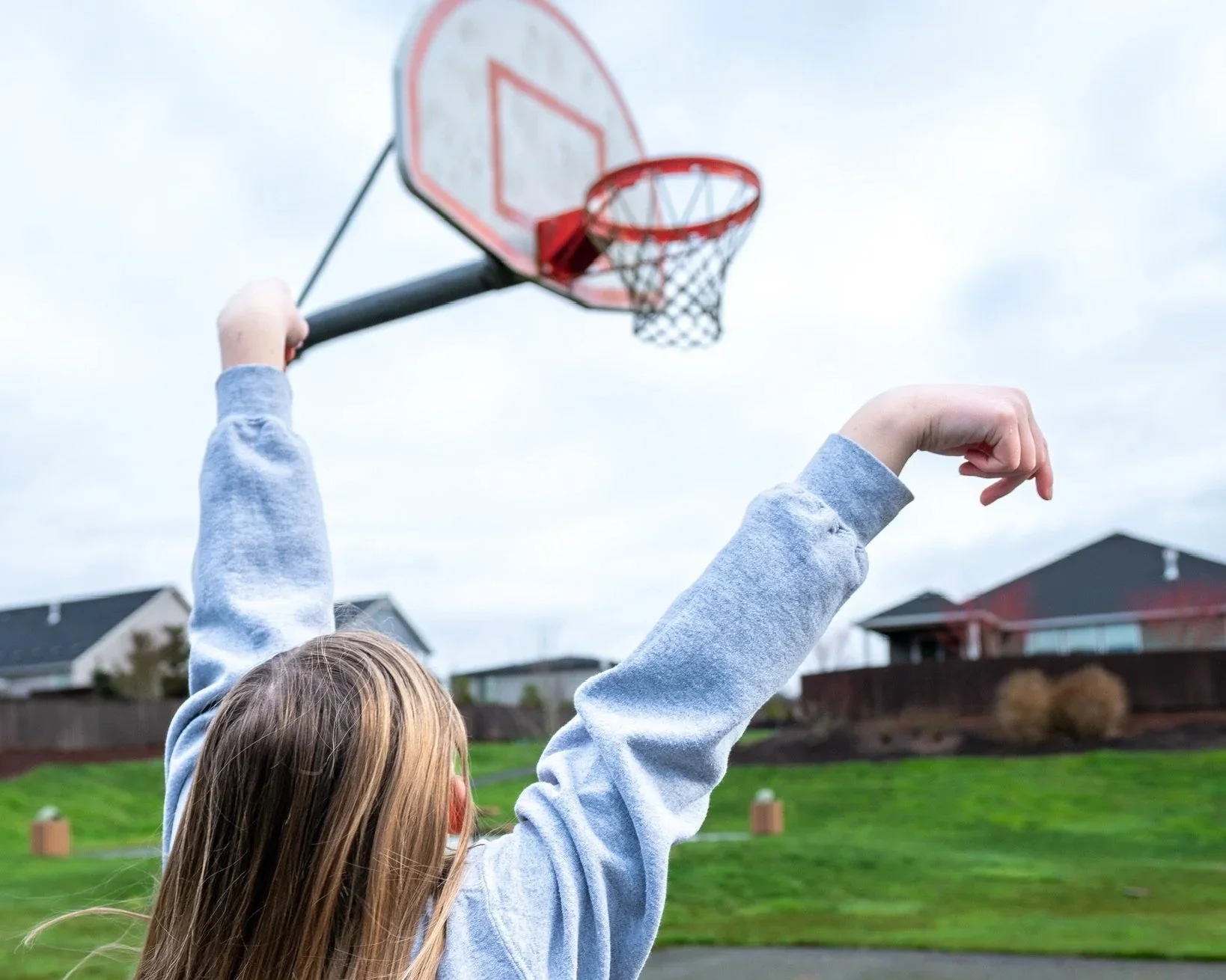 A girl with long hair in a grey hoodie is playing basketball outdoors near suburban houses on a cloudy day.