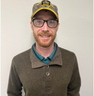 A man with glasses, a beard, and a mustache, wearing a yellow and black cap, a green shirt under a brown sweater, smiling against a plain background.