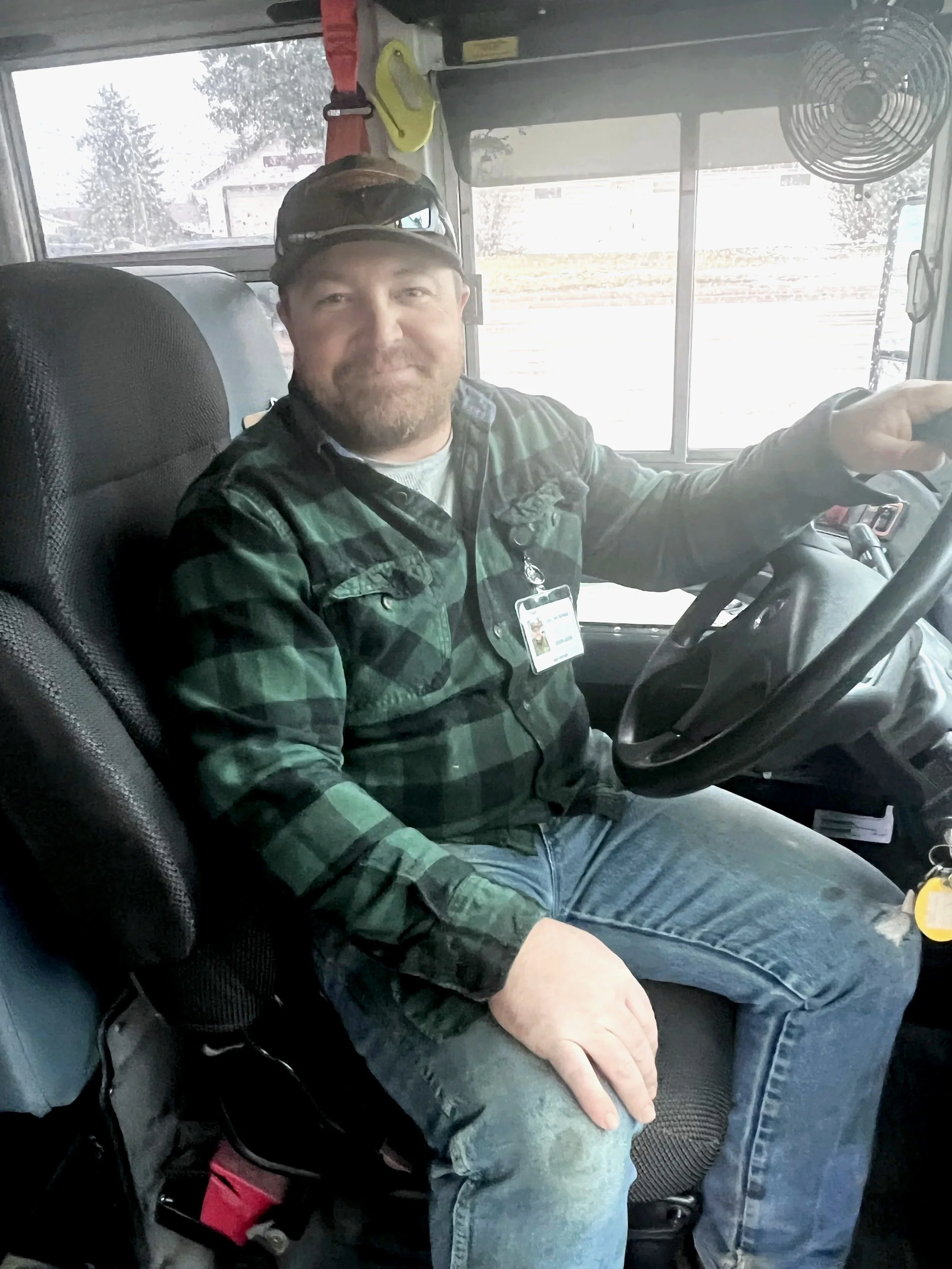 A man sitting in the driver's seat of a vehicle, smiling, wearing a green plaid shirt, jeans, and a hat, inside a vehicle with a dashboard and a window behind him.